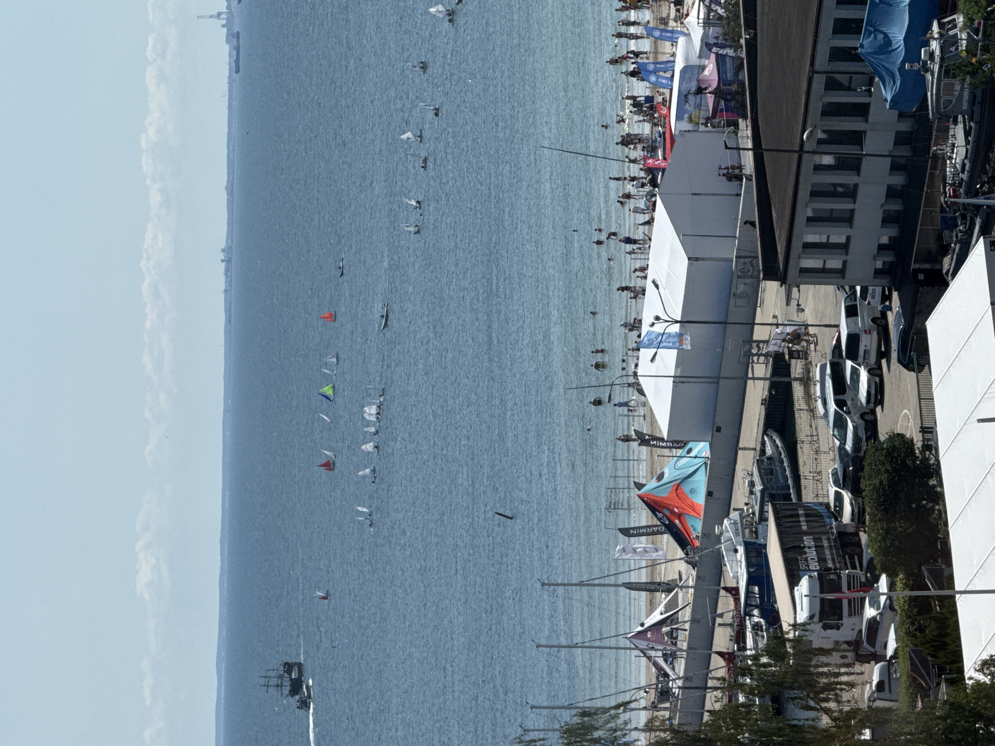 The image captures a view of the sea and a populated beach area in Gdynia, Poland. On the water, a large sailing ship, possibly a replica, is visible on the left, accompanied by a smaller boat. Numerous smaller sailboats are scattered across the sea, with some participating in what appears to be a regatta, indicated by their sails in various colors. The beach itself is crowded with people, suggesting a warm, sunny day. Several white tents are set up on the sand, some serving as vendor stalls or event shelters, with flags and signage visible. In the foreground, a harbor area is depicted, with boats docked and a large truck bearing the logo "SPORT evolution" parked on the quay. The overall scene is a lively maritime and beach environment.