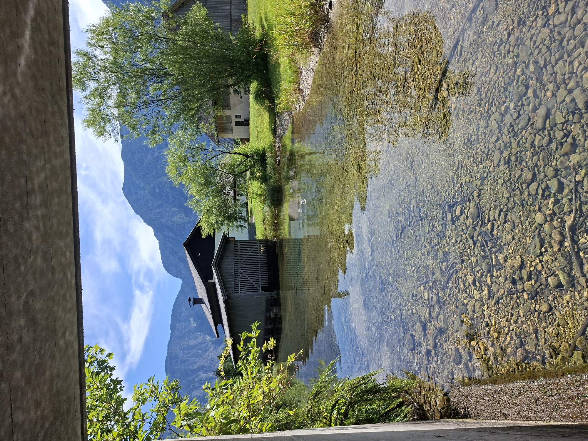 This is a photograph taken from a bridge looking out over a clear, shallow body of water with a rocky bottom. In the foreground, the edge of the concrete bridge structure is visible on the left, with lush green foliage hanging over the water. The water reflects the sky and surrounding trees.
Across the water, there is a wooden boathouse with a dark roof and a large, open doorway. Behind the boathouse, there are trees and a building with a light-colored facade and a dark roof. Further in the background, large, tree-covered mountains rise under a bright blue sky with scattered white clouds. The scene depicts a serene, natural environment likely in Obertraun, Austria, given the architectural style and mountainous landscape typical of the Salzkammergut region. There are no people or animals visible in the image.