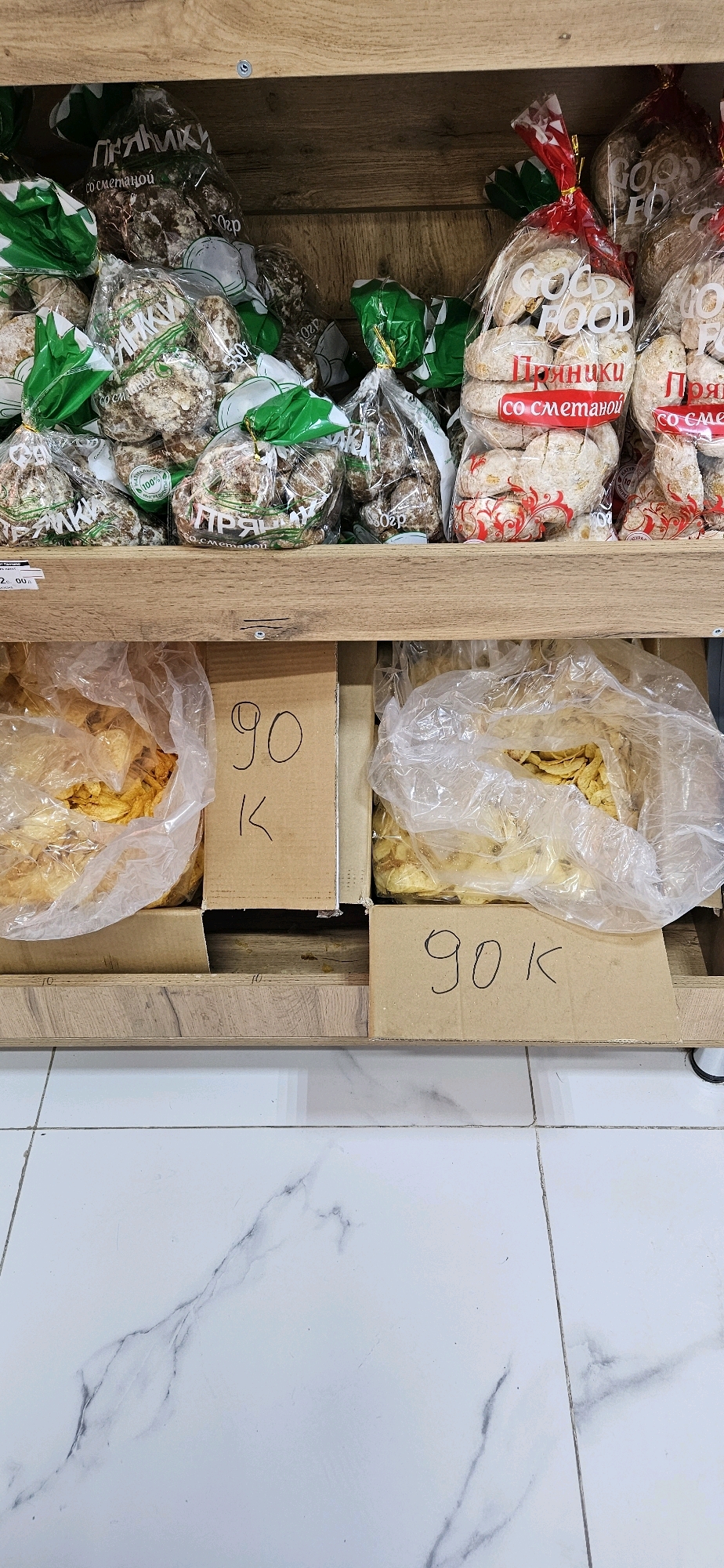 A high-angle, eye-level shot captures a wooden shelf filled with various packaged food items, primarily baked goods, on the top shelf and what appear to be snacks or chips on the bottom shelf. The top shelf displays an assortment of cookies and pastries in clear plastic bags, some with green ties and labels in what appears to be Cyrillic script. Others are in bags with red and white branding that says "GOOD FOOD". The bottom shelf holds two large, clear plastic bags filled with yellow, irregular-shaped food items, possibly potato chips or dried fruit. Placed in front of these bags are two pieces of cardboard, each with handwritten numbers. The left cardboard shows "90 K", and the right cardboard also displays "90 K". The shelf is made of wood with visible grain. The floor below the shelf is tiled with a white marble pattern. The overall scene suggests a retail environment, possibly a small grocery store or market stall.