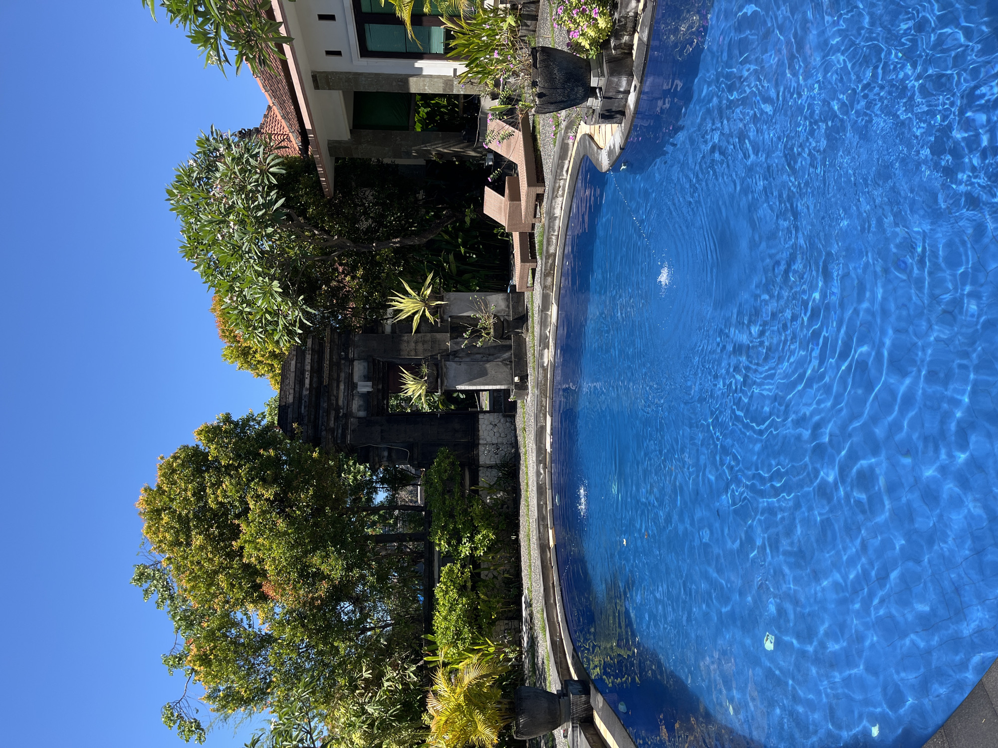 The image depicts a section of a swimming pool in Kuta Selatan, Indonesia. The pool is filled with clear blue water, rippling slightly. Adjacent to the pool is a building with a red-tiled roof and a facade adorned with lush tropical plants and trees. A dark stone structure, possibly an ornamental gate or wall, is present between the greenery and the pool area. The sky is clear and blue. There are no people visible.