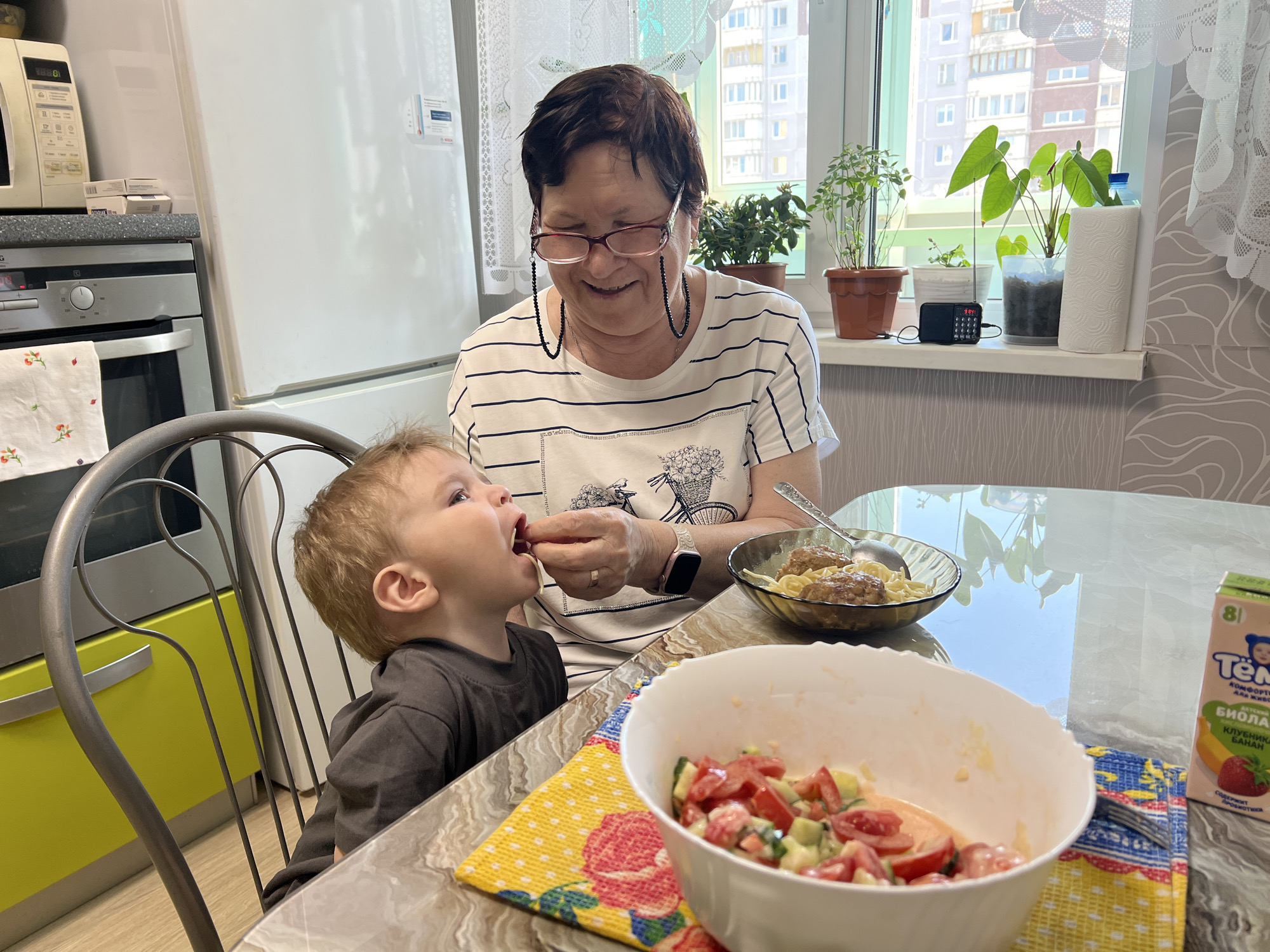 The image depicts an indoor scene in a well-lit kitchen in Perm, Russia, during what appears to be a bright, sunny day. An elderly woman with short dark hair and glasses, wearing a striped t-shirt with a floral basket graphic, is seated at a glass-top dining table. She is smiling and gently feeding a young child, who has light brown hair and is looking up at her with an open mouth, seated in a high chair or booster seat.
The kitchen is modern and features white and stainless steel appliances. To the left, a white Samsung microwave displays "08:50" on its digital clock, positioned above a stainless steel AEG oven with control knobs. A tall, white Bosch refrigerator stands beside the oven. Bright yellow lower cupboards are visible beneath the countertop.
On the dining table, there are two bowls of food: one glass bowl contains what looks like spaghetti with meatballs, and a white bowl holds a fresh salad of chopped tomatoes and cucumbers. A colorful, patterned napkin is placed under the salad bowl. A small carton of "Тёма БИОЛАКТ" (Tёma Biolact) "КЛУБНИКА БАНАН" (Strawberry Banana) with an "8+" (indicating suitable for 8 months and older) label is also on the table. The woman wears a white smartwatch on her left wrist.
Behind the woman, a large window offers a view of several multi-story residential buildings, suggesting an urban setting. On the windowsill, there are multiple potted plants, a small black radio displaying "3:04", and a roll of paper towels. The natural light flooding the room and the tender interaction between the woman and child create a warm and caring atmosphere.