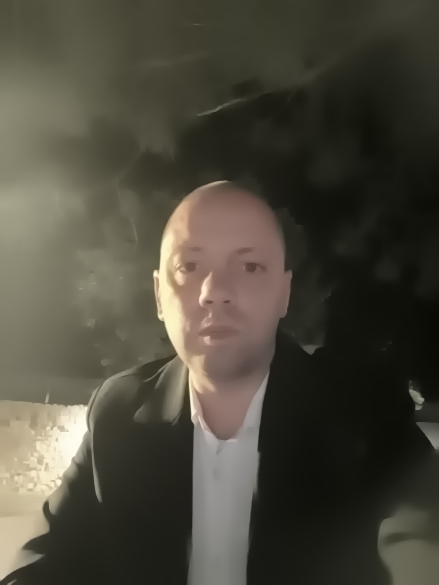 The image is a medium shot of a bald man wearing a white shirt and a black blazer. The background appears to be a dark, blurry outdoor setting, possibly at night. The lighting is dim, and the location is suggested to be somewhere in Нареченски бани, Bulgaria. The man is looking directly at the camera with a neutral expression.