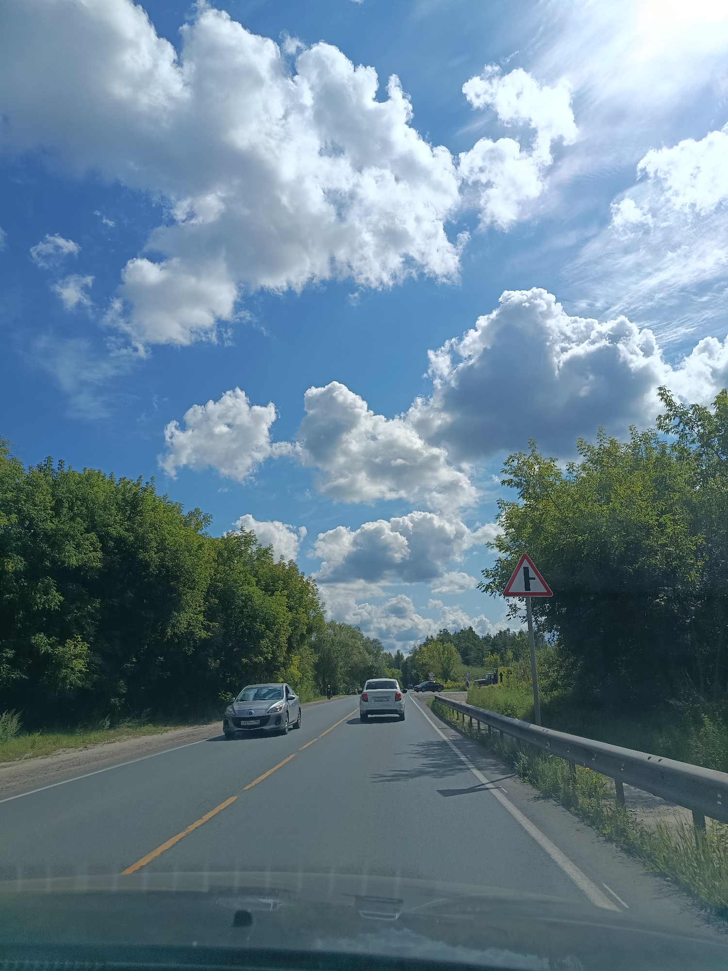 The image captures a view from inside a vehicle traveling on a two-lane asphalt road lined with lush green trees. The sky is a vibrant blue, dotted with fluffy white clouds. A silver sedan is visible in the left lane, and a white SUV is in the right lane ahead. To the right of the road, a metallic guardrail runs alongside, and a triangular traffic sign with a black 'T' symbol is mounted on a pole. In the distance, the road curves, and more trees and vegetation are visible. The scene suggests a sunny day on a rural or suburban road. No specific landmarks are identifiable to pinpoint a precise location within Orekhovo-Zuyevo, Russia.