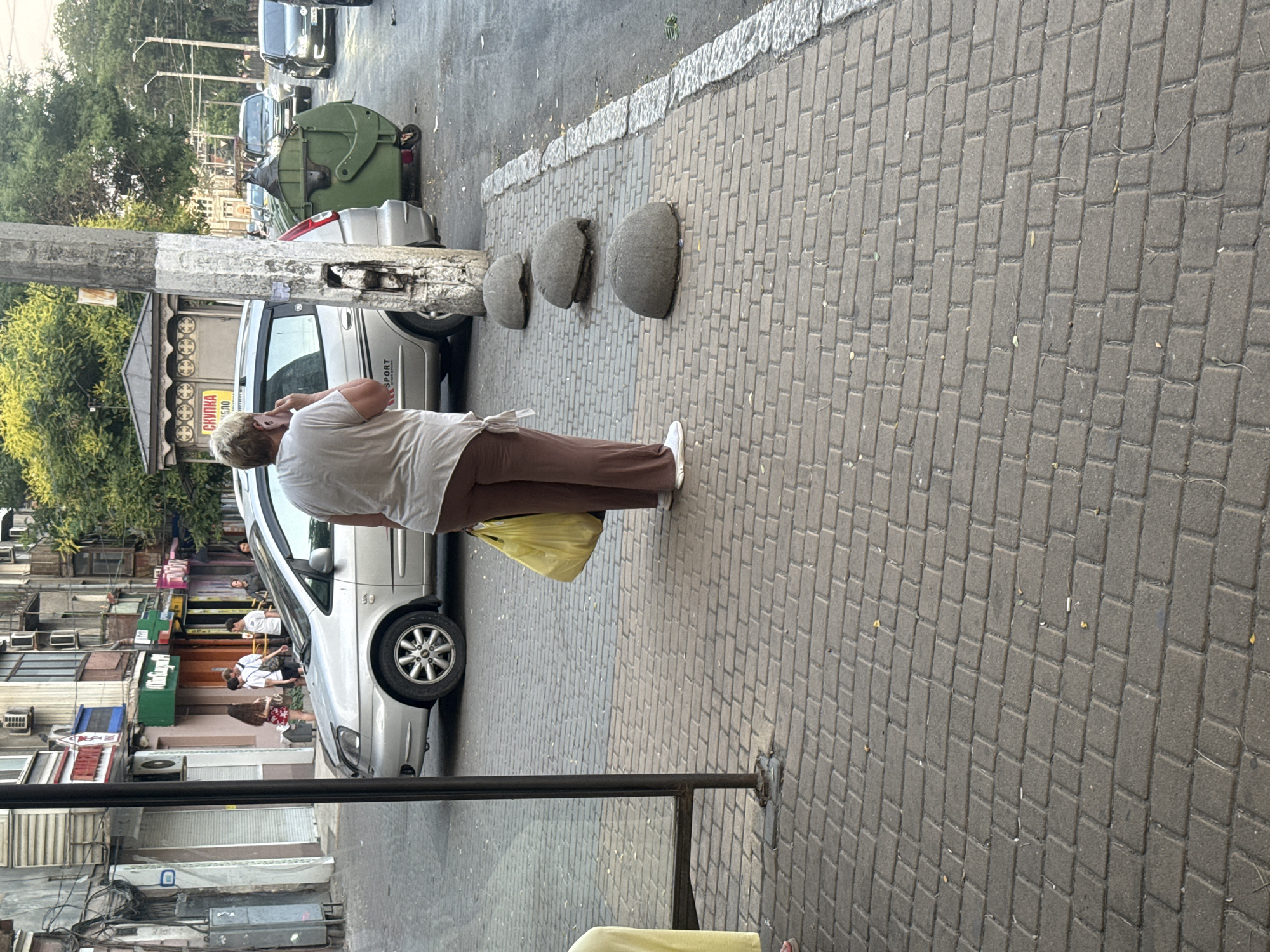 In the city of Odesa, Ukraine, on a paved street, a woman stands at a bus stop, talking on her phone. She is wearing a light-colored t-shirt and brown pants, and is holding a yellow plastic bag. Behind her, a silver car is parked next to a large green dumpster. The street is lined with buildings, some of which have shops on the ground floor. In the background, other cars are visible, and trees line the street. The ground is paved with brick-like stones, and there are several round, grey bollards along the sidewalk. The overall scene depicts a typical urban streetscape in Odesa.