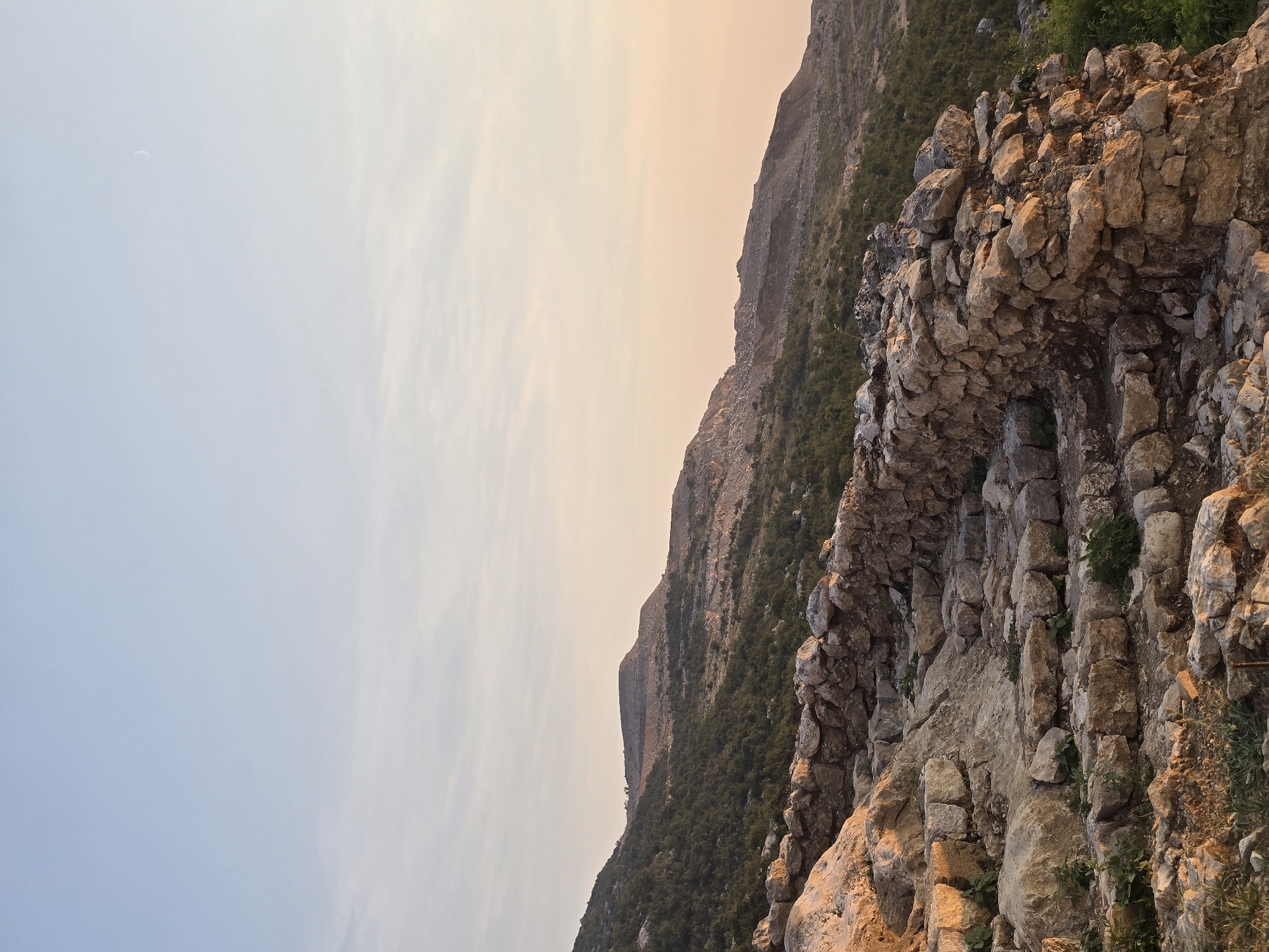 An outdoor, eye-level, medium shot captures an ancient stone structure, possibly ruins of a fortress or castle, set against a backdrop of mountainous terrain. The stone structure, constructed from irregular, rough-hewn rocks of varying sizes, forms a wall and a series of steps ascending towards the right of the frame. The rocks are a mixture of earthy tones, with some appearing lighter and sun-bleached, while others are darker, possibly due to moisture or shadowing. Green vegetation, appearing as small bushes and grasses, sprouts from between the stones and along the path, indicating the passage of time and the encroachment of nature.

The landscape beyond the stone structure is characterized by rolling hills covered in dense green foliage, transitioning into rocky, barren slopes further in the distance. The uppermost portions of the mountains are indistinct, blending into the sky.

The sky occupies the upper half of the image and exhibits a gradient of soft colors characteristic of dawn or dusk. The lower part of the sky is a pale, warm peach hue, which gradually transitions into a clear, pale blue at the top. A faint, thin crescent moon is visible in the upper right quadrant of the sky.

The lighting suggests it is either early morning or late evening, with the sun casting a warm glow on the stone structure, highlighting its textures and contours. The overall impression is one of historical significance and natural beauty. The scene is devoid of any human presence or activity.