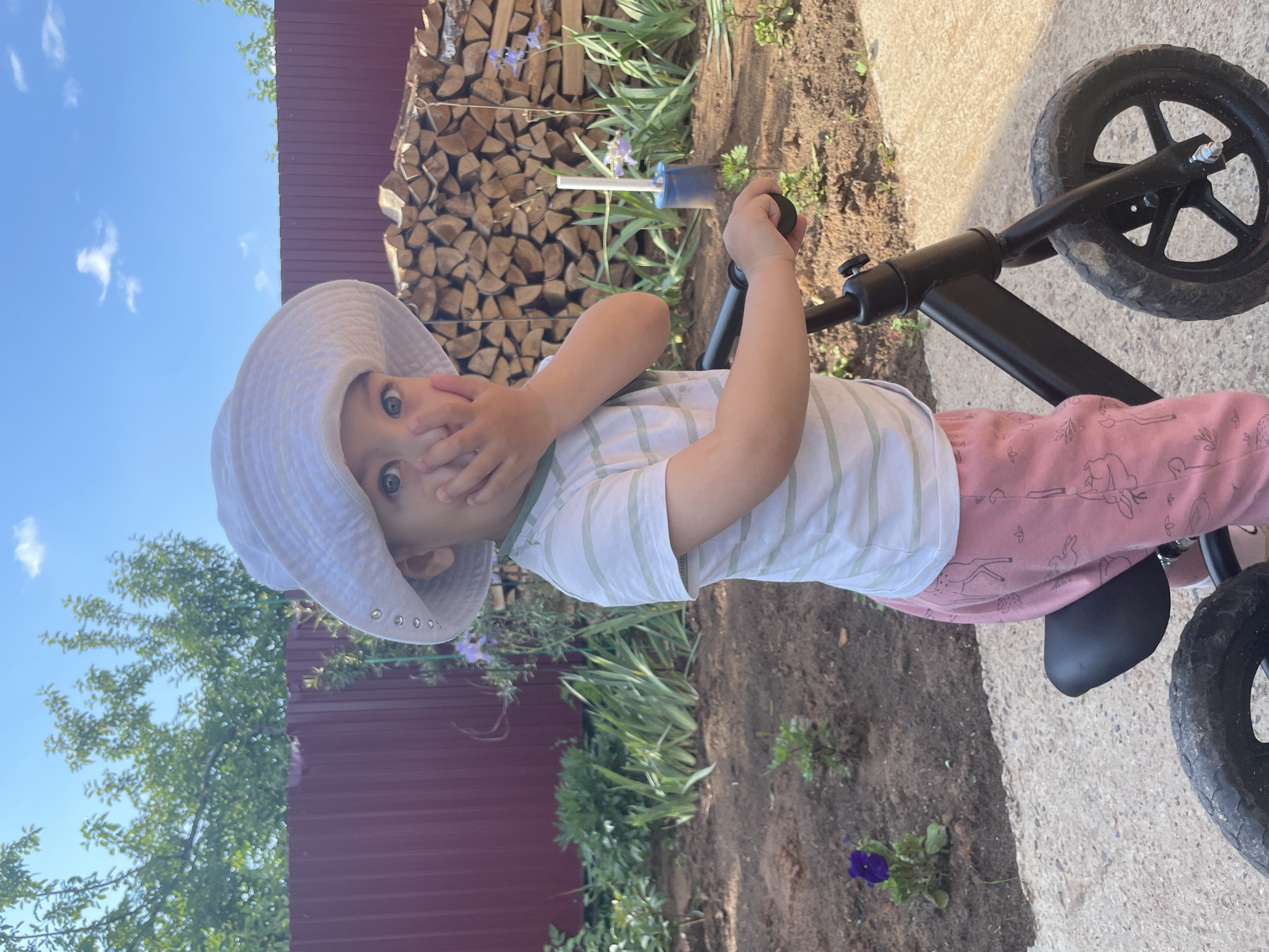 A young child wearing a white sun hat, a striped t-shirt, and pink pants with deer patterns is riding a black balance bike outdoors in what appears to be a backyard. The child is looking at the camera with their hand covering their mouth, suggesting surprise or shyness. The background features a red fence, a pile of firewood, plants, and a blue sky with some clouds. The ground is a mix of dirt and a paved surface. The scene is likely set in Zav'yalovo, Russia, given the context.