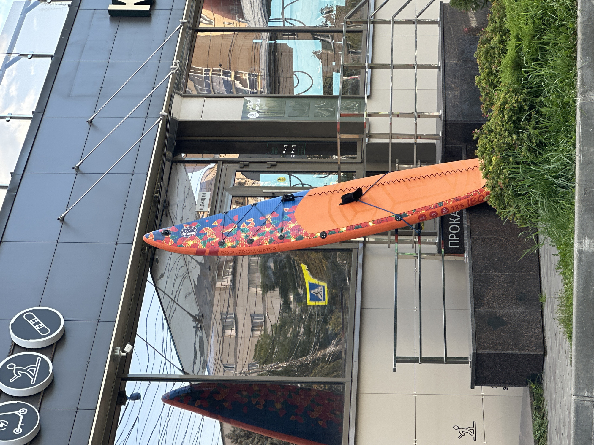 A vibrantly colored paddleboard, leaning upright against a building's facade, is the central subject of this outdoor urban scene. The paddleboard displays a complex pattern of orange, blue, and red, with floral and string-like motifs. It is positioned in front of a large, reflective glass window, which mirrors the surrounding environment, including buildings and power lines. Above the window, three circular signs are mounted on the building's dark, tiled exterior. These signs feature simple white icons: one of a scooter, another of a person on a snowboard, and a third of a snowboard. Part of a building's name, with the letter "K" visible, is also present on the facade. Below the window, a short dark stone planter box is topped with a sleek, metallic railing. Lush green bushes and grass grow around the planter box and in front of the building. The ground in the foreground is paved, with a concrete curb visible. The overall impression is that of a commercial establishment, possibly a sports or outdoor recreation store, given the prominent display of the paddleboard and the icons on the signage. The lighting suggests it is daytime, with diffused sunlight.