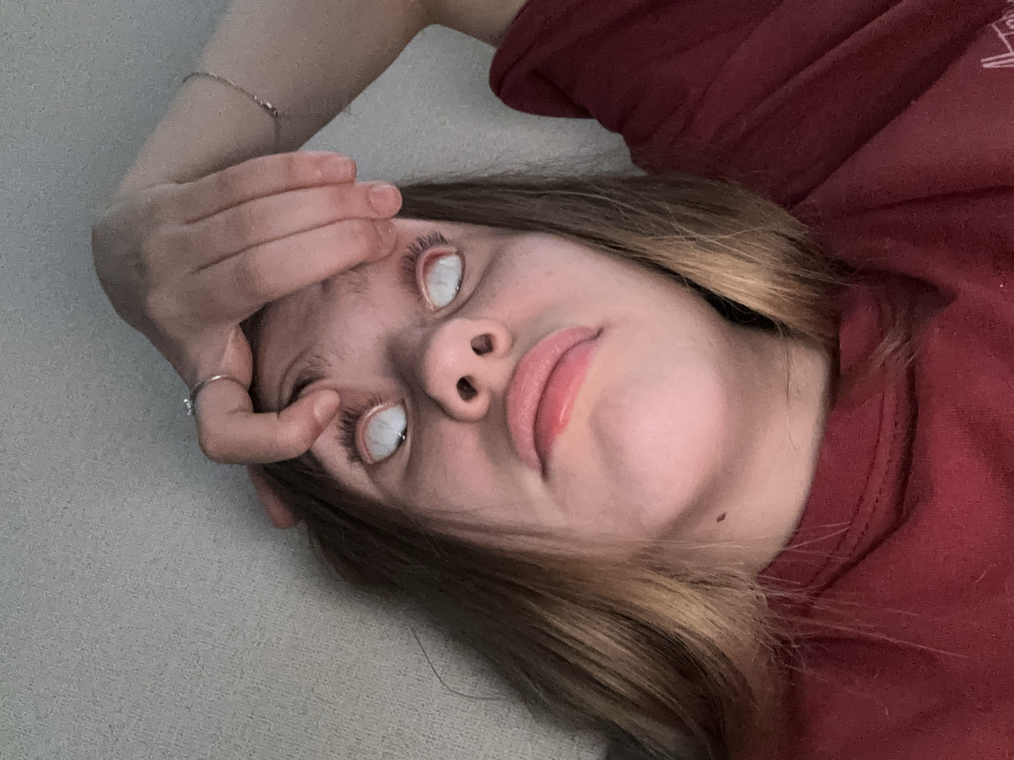 A young female is lying down, holding up her upper eyelids with her right hand. Her eyes are open wide and appear to have white scleras and no visible irises or pupils. Her mouth is slightly open, and she is looking upwards. She is wearing a red t-shirt. A ring is visible on her right ring finger, and a thin bracelet is on her right wrist. The background is a textured, light-colored surface, possibly a wall or sofa. There are no discernible city landmarks or features that would indicate a specific location in Vsevolozhsk, Russia.