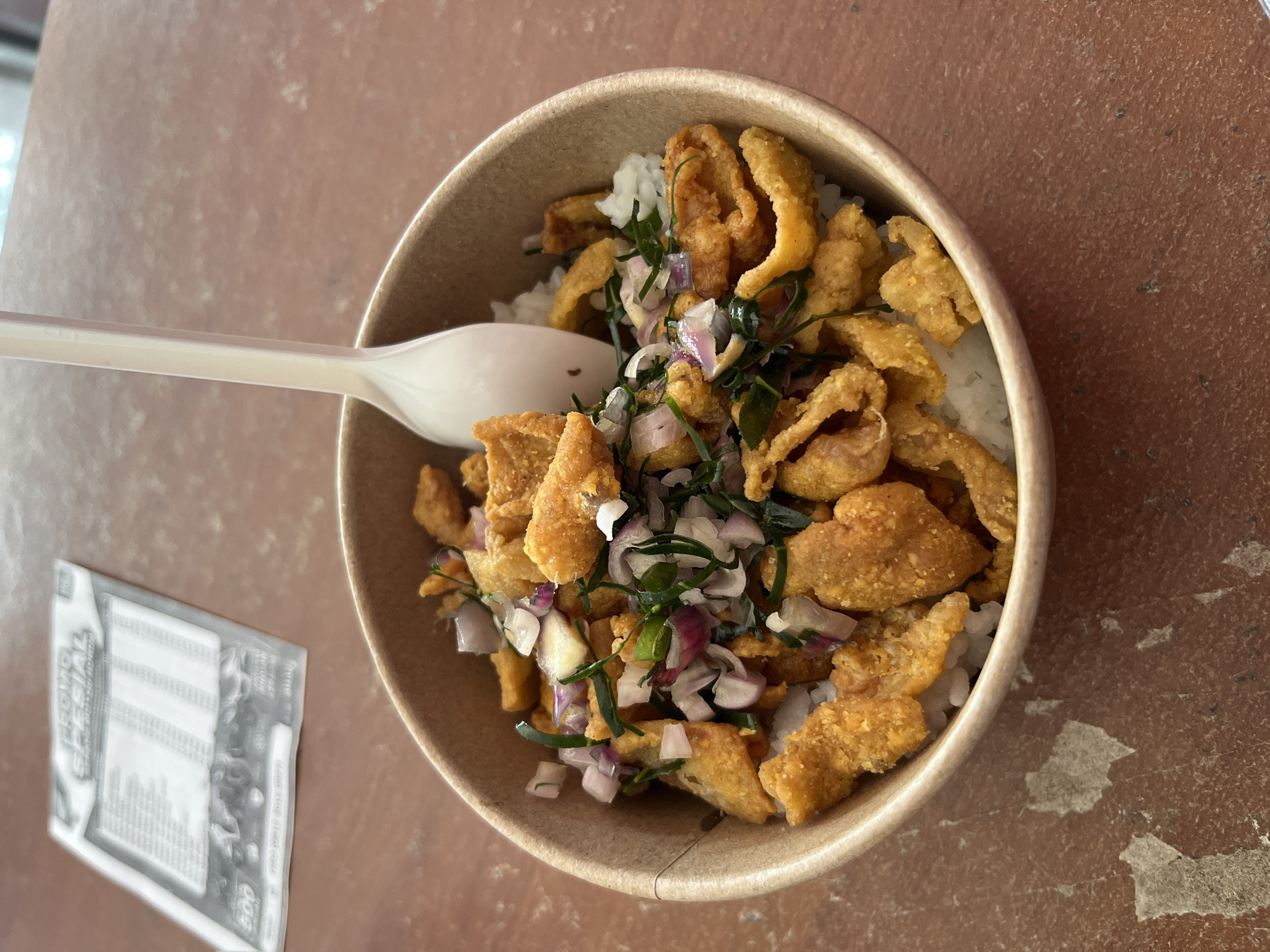 A bowl of white rice topped with crispy fried chicken pieces, chopped red onions, and green herbs is presented on a brown textured surface. A white plastic spoon rests in the bowl. In the background, a blurred menu or flyer displays text and a grid. This scene is located in Boja, Indonesia.