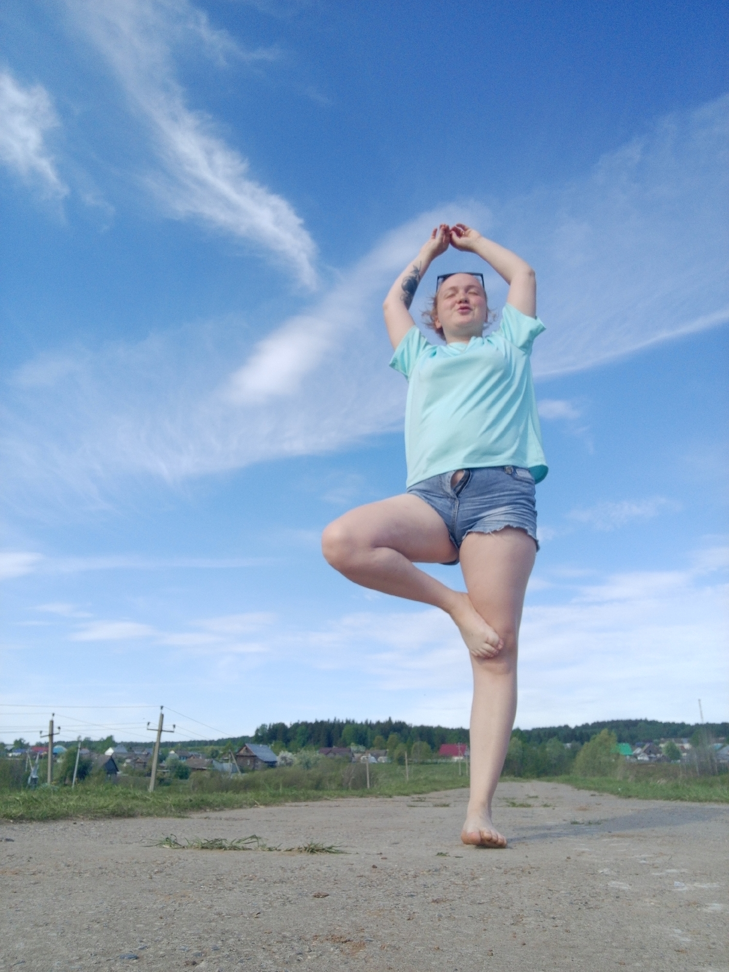 The image shows a woman doing a tree pose in an outdoor environment. She is wearing a light blue t-shirt and jean shorts, and she is barefoot. Her arms are raised above her head, and her hands are clasped together. She has sunglasses on top of her head. The background consists of a blue sky with scattered clouds, a dirt road, and some trees and buildings in the distance. The location appears to be Уральское, Russia, based on the presence of typical rural Russian structures, such as wooden houses with colorful roofs and electrical poles.