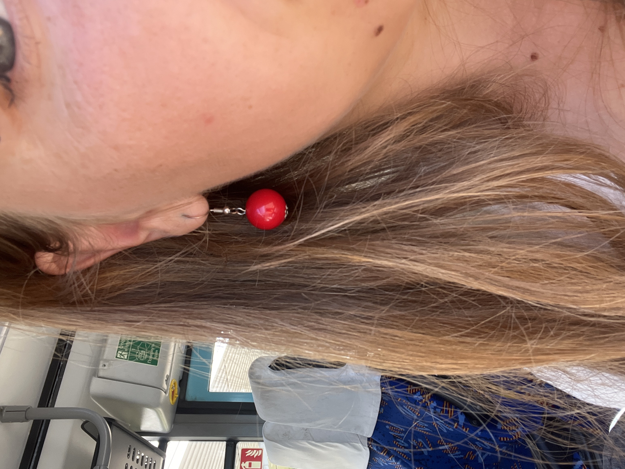 A close-up, medium-shot photograph captures a woman's right ear and cheek, adorned with a dangling red earring. Her long, blonde-brown hair cascades around her face, partially obscuring the background. The environment suggests the interior of a public transport vehicle, possibly a bus, with hints of seating covered in a blue and orange patterned fabric. In the upper left corner, a white, rectangular emergency sign with green text and graphics is visible, indicating this image was taken in Myadzyel, Belarus. The lighting is bright, suggesting daytime.
