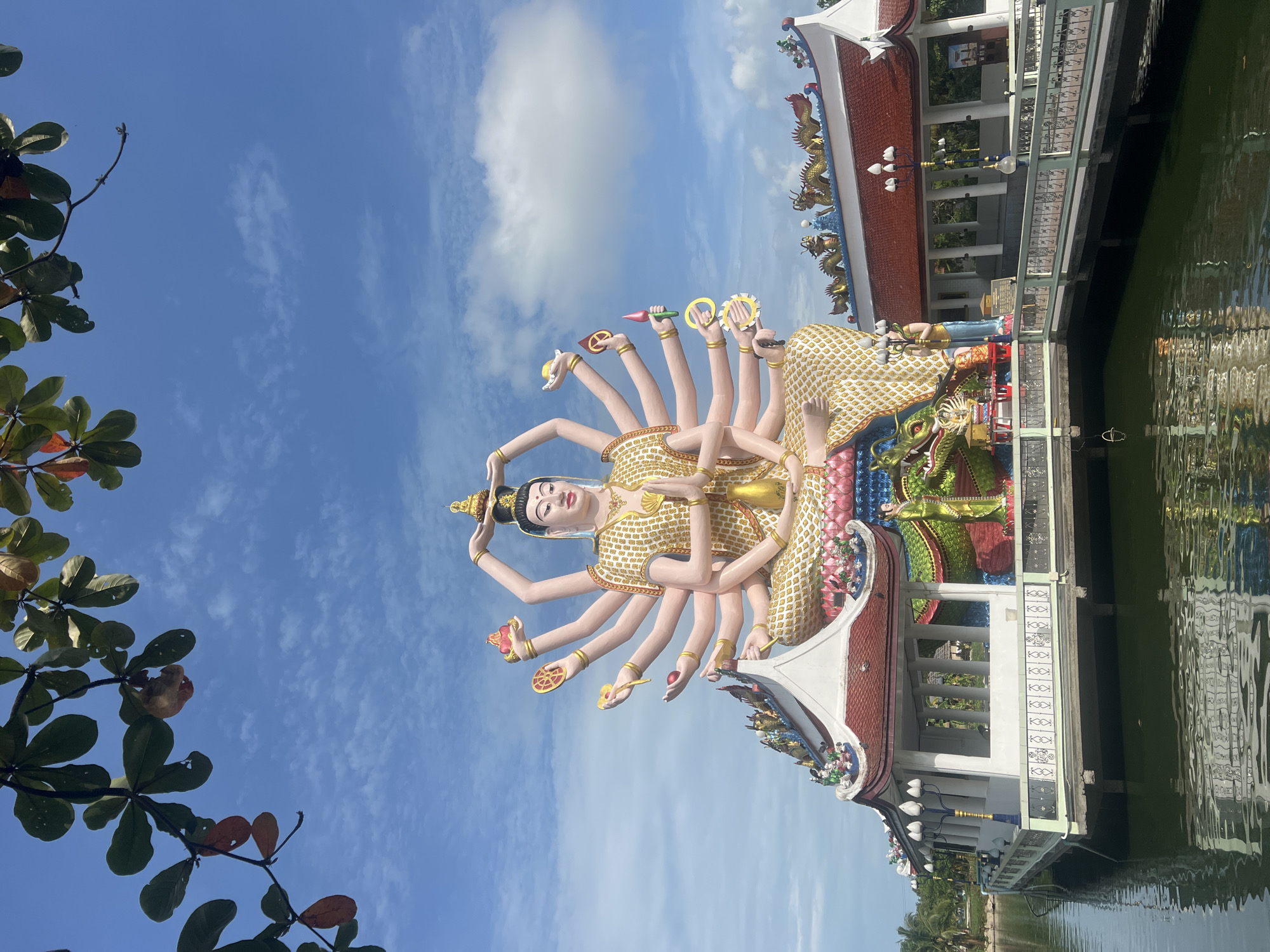 The image depicts a large, colorful statue of Guanyin with multiple arms, located at Wat Plai Laem in Baan Plai Laem, Thailand. The statue is adorned in a gold and white patterned robe and stands on a platform with an ornate roof decorated with dragons. A building with similar architectural style is seen adjacent to the statue, partially overhanging a body of water, possibly a pond or lake. Reflections of the building and surrounding structures are visible in the water. The sky is blue with scattered clouds, and the branches of a tree with green and reddish-brown leaves are visible in the lower-left corner.