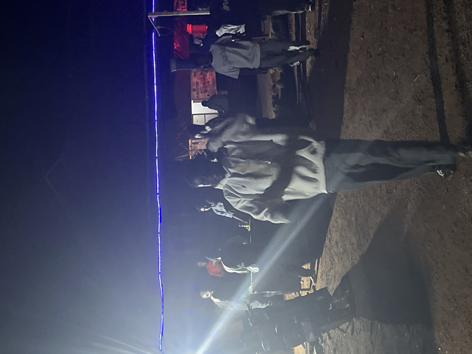 In the city of Durumi, Nigeria, at night, a group of people are gathered outdoors. A bright light source, possibly a spotlight or flash, illuminates the left side of the scene, casting lens flare. The gathering appears to be informal, with several individuals standing around a table in the background. One person in the foreground is wearing a light-colored hooded sweatshirt and dark pants, walking towards the camera. Blue LED lights are strung across the top of the scene, indicating some form of decoration or illumination for an event. The ground is dirt and uneven. In the background, there are structures that suggest a building or shelter, and some vehicles are partially visible on the left. The overall atmosphere suggests a social gathering or event taking place outdoors at night.