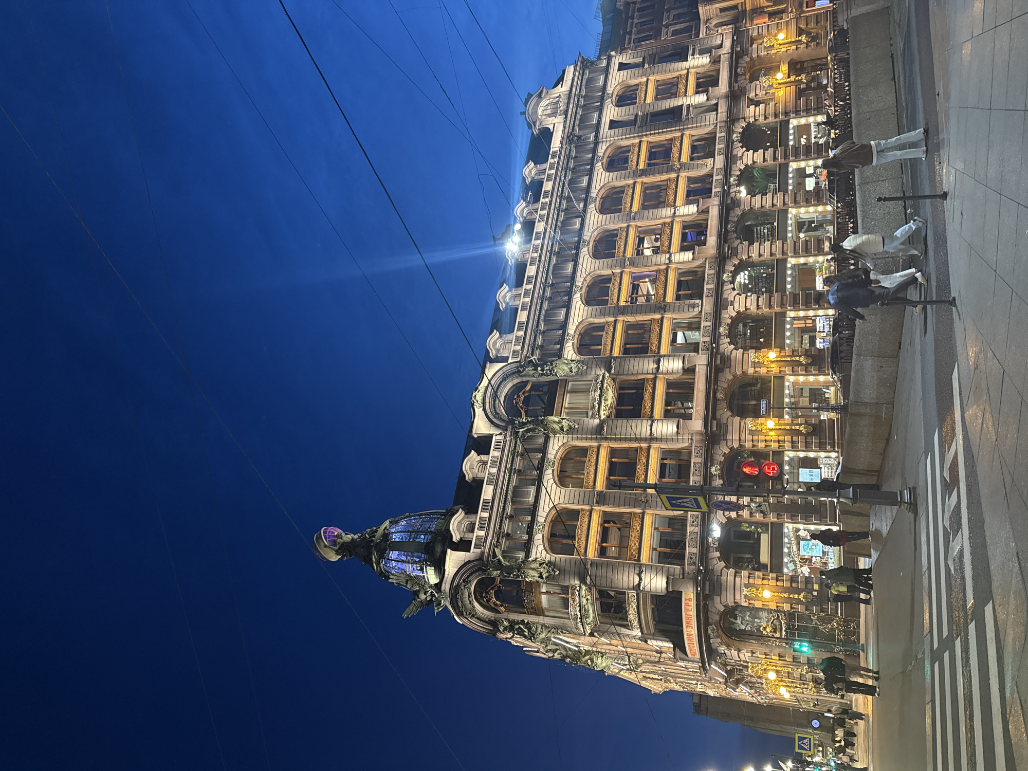 The image shows the Singer House (Dom Knigi), a historic building on Nevsky Prospekt in Saint Petersburg, Russia, at dusk. The building features an ornate facade with arched windows, decorative sculptures, and a prominent glass dome topped with an eagle statue. The dome is illuminated with purple and blue lights.
Streetlights and interior lights from the shops on the ground floor illuminate the building's facade and the surrounding street. The sky is a deep blue, indicating the transition from day to night.
Several people are visible on the sidewalks and crossing the street at a crosswalk. Some are walking individually, while others are in small groups. Traffic signals and street signs are also present. Power lines are strung across the sky, a common sight in urban environments. The overall scene depicts a vibrant city street at twilight, with the Singer House as the central architectural landmark.
