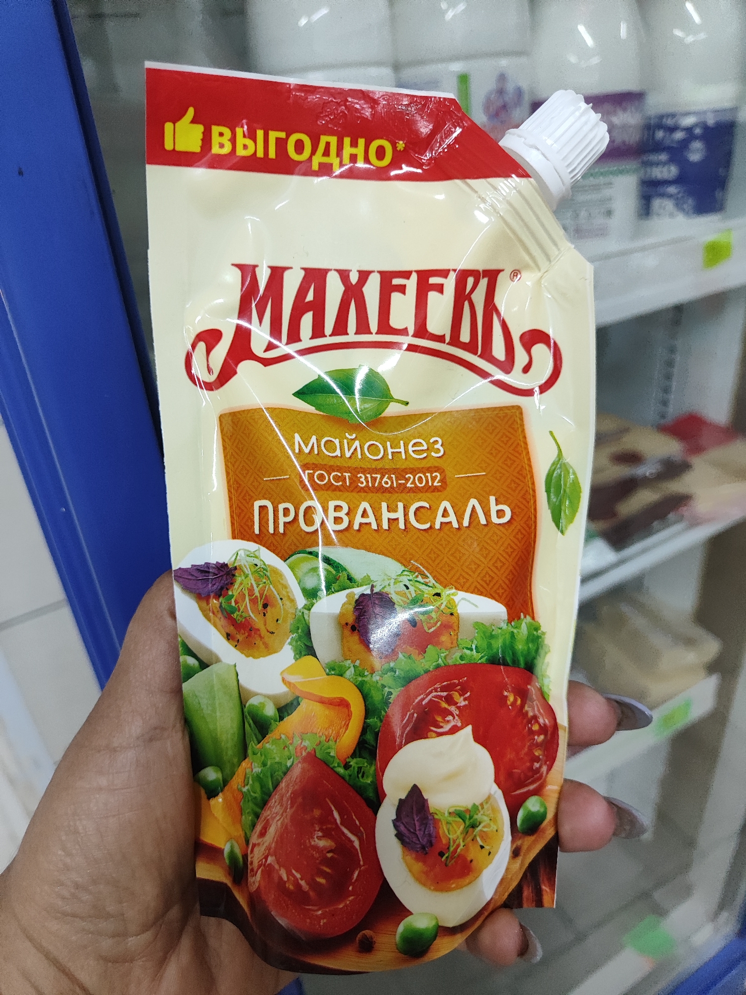 A person's hand is holding a pouch of mayonnaise in front of a shelf in what appears to be a store. The pouch is mostly yellow with red accents. The text on the pouch is in Russian and includes "МАХЕЕВЪ" in large, stylized letters, and "МАЙОНЕЗ" and "ПРОВАНСАЛЬ" in smaller text. The image on the pouch depicts a salad with sliced tomatoes, hard-boiled eggs, lettuce, and olives. The background shows shelves with other products, some of which are blurred. The lighting is bright, suggesting an indoor setting.