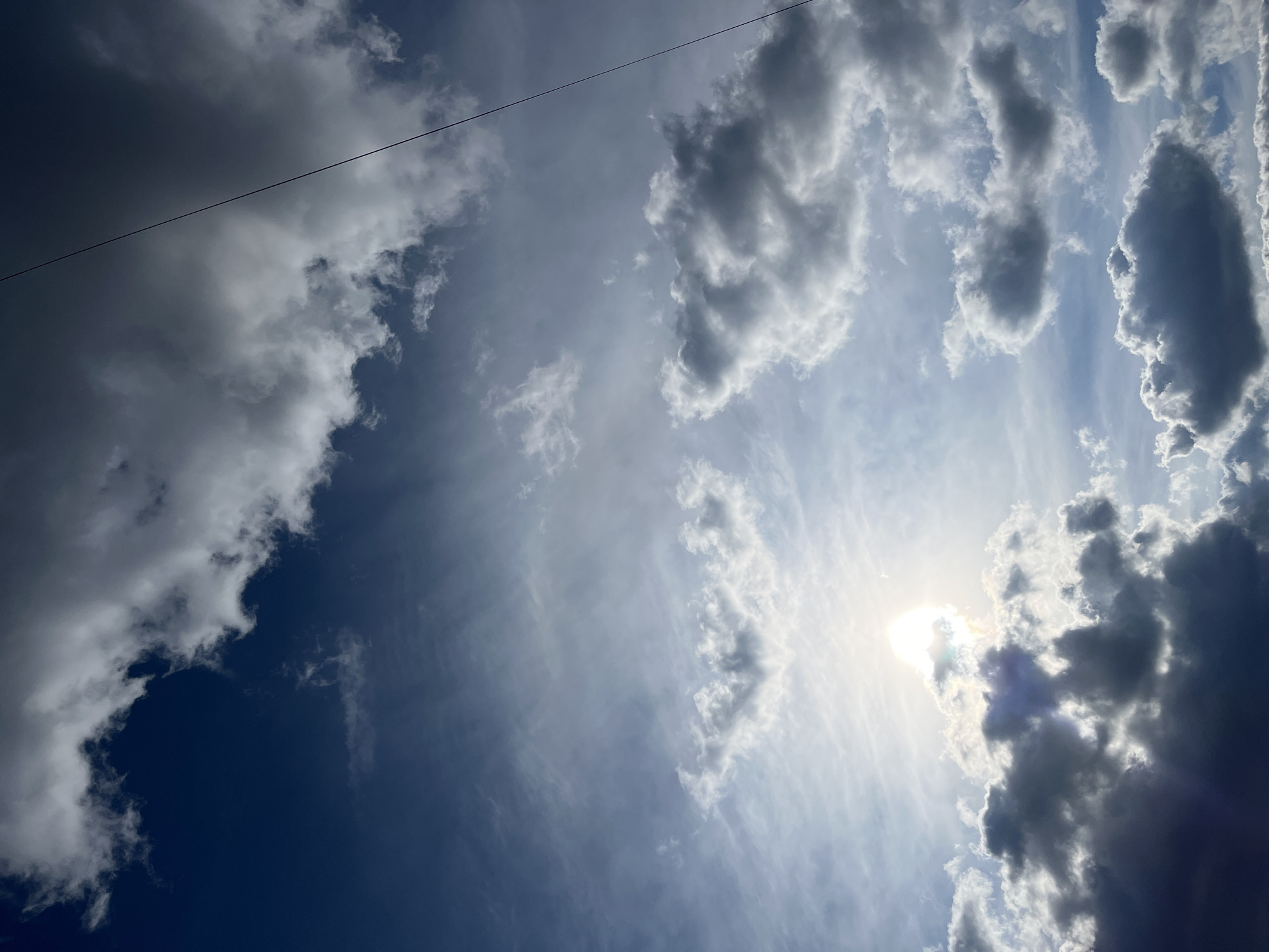 The image shows a daytime sky with scattered cumulus clouds. The sun is partially obscured by clouds in the lower right, creating a bright area. A dark blue portion of the sky is visible in the lower left. A power line stretches across the top left of the frame. The image does not provide specific details indicating the location in the city of Klin, Russia, nor does it include any people, subjects, objects, actions, or interactions.