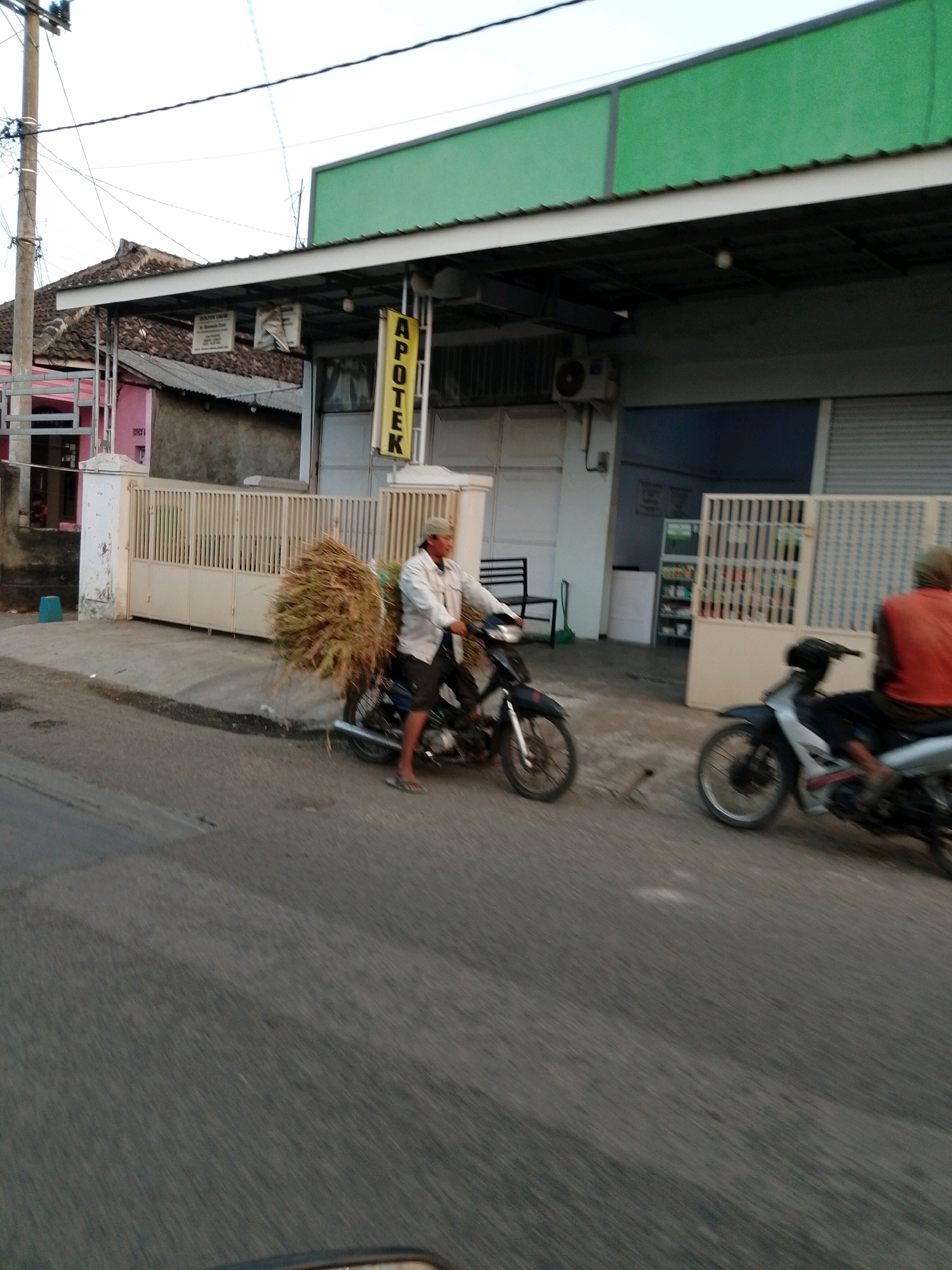 A motorcycle driver wearing a light-colored jacket and dark shorts, with a baseball cap on his head, is riding a motorcycle. He is transporting a large bundle of dried grass or straw strapped to the back of his motorcycle. The motorcycle is positioned in front of a beige metal gate, which is part of a building with a green overhang. A yellow sign with the word "APOTEK" (Indonesian for "pharmacy") is visible on the building. Another motorcycle with a person wearing an orange shirt is further down the road to the right. The scene appears to be a street in the city of Pasuruan, Indonesia. The road surface is asphalt, and there are electrical poles and wires overhead. The lighting suggests it is either early morning or late afternoon.