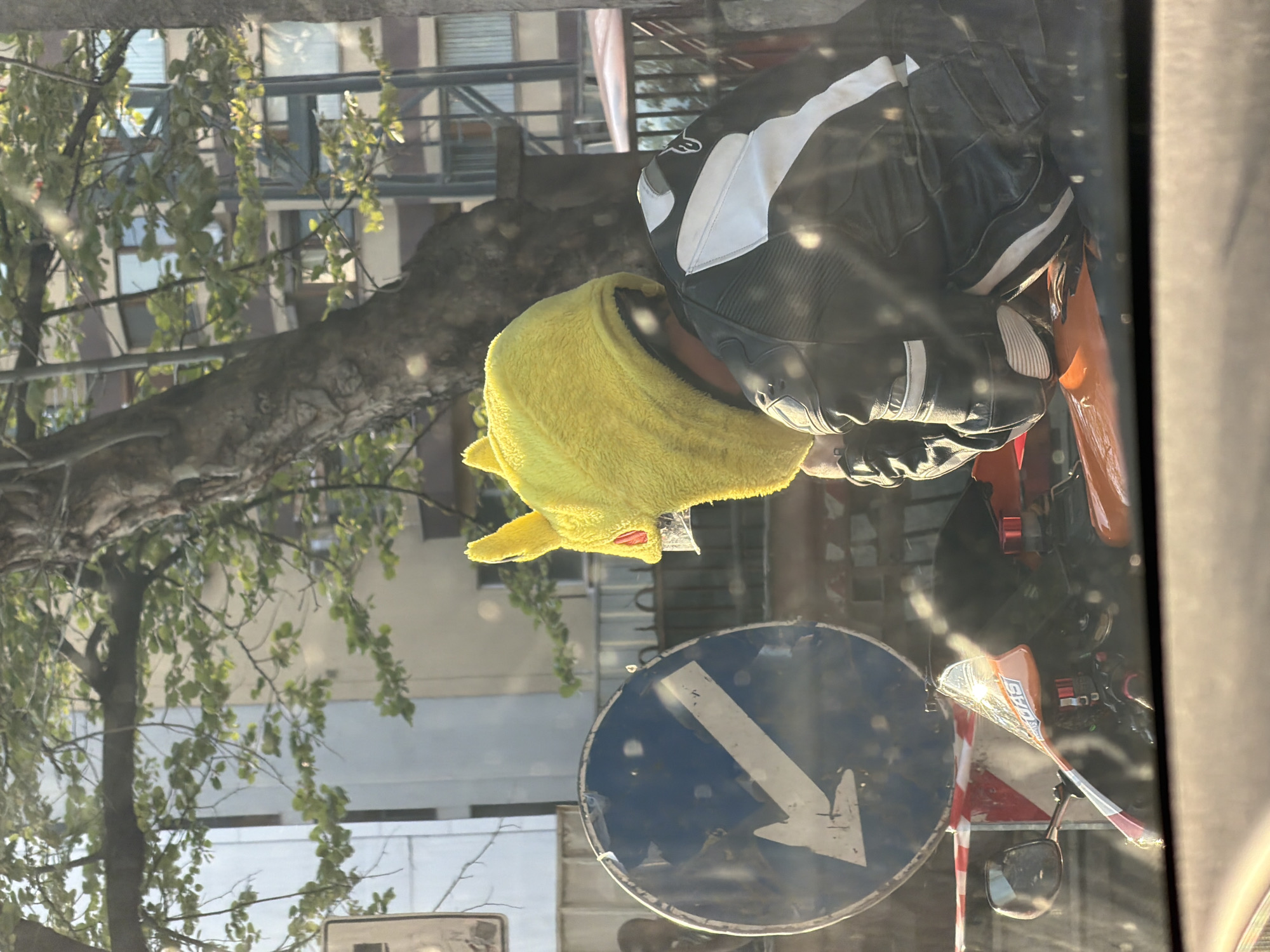 The image shows a person on a motorcycle wearing a yellow hood. The person is wearing black and white riding gear. A blue road sign with a white arrow pointing downwards is visible in the foreground. The background shows a tree with green leaves and buildings, possibly in a city.