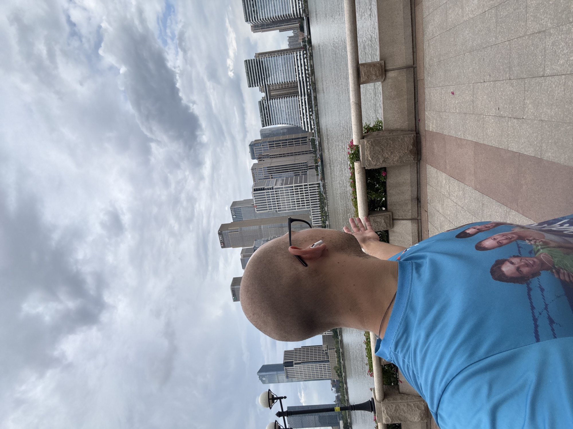 The image shows a man from the back, with his head tilted upwards and to the side. He is wearing a blue t-shirt with a graphic print on it featuring three faces. The man has a shaved head and is wearing glasses perched on his head, with one earbud in. He is raising his right hand, palm facing outwards, as if waving.
The man is standing on a paved walkway next to a body of water, likely a river or bay. Across the water, a city skyline is visible, characterized by numerous tall, modern buildings. The sky is cloudy and overcast, suggesting it is daytime but not necessarily sunny. The walkway has a stone railing and planters with some greenery and pink flowers.
The scene appears to be in a developed urban area, with the buildings suggesting a bustling city. The overall mood is casual, possibly indicating the man is enjoying a view or interacting with someone. The context of Tianhe District, China, places this scene within a well-known commercial and residential hub. There is no discernible text in the image.