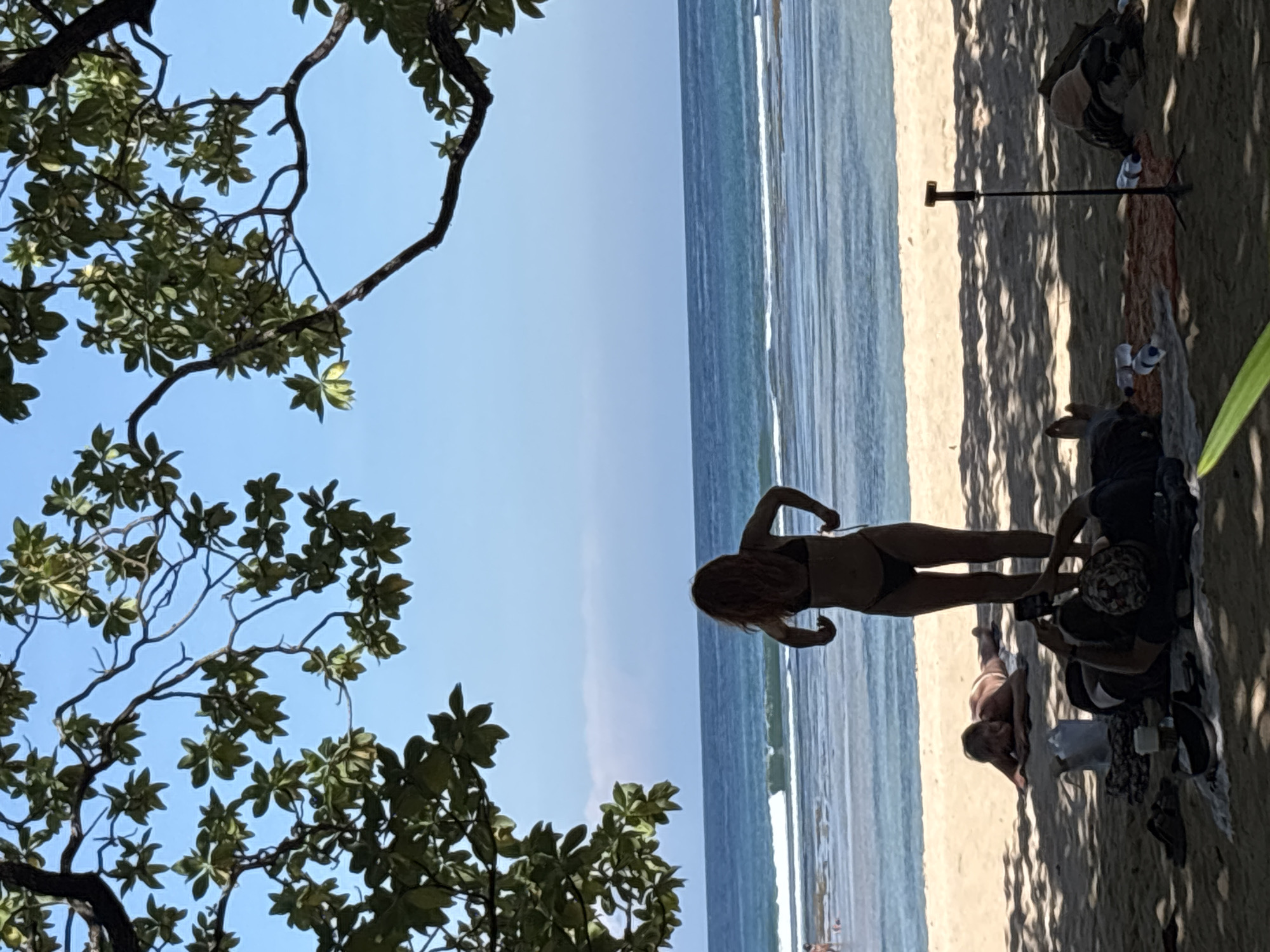 The image depicts a beach scene in Kuta Selatan, Indonesia. The horizon line is nearly parallel with the top and bottom edges of the frame. The sky is clear blue. The ocean has gentle waves. In the middle of the frame, a woman in a black bikini stands on the sand, facing the ocean. To the right, under the shade of what is likely a tree, a group of people are sitting on the sand. One person appears to be taking a photo with their phone. A stand is visible, and some bags and other items are scattered around. The left side of the frame is dominated by tree branches and leaves. The photo is rotated 90 degrees clockwise.