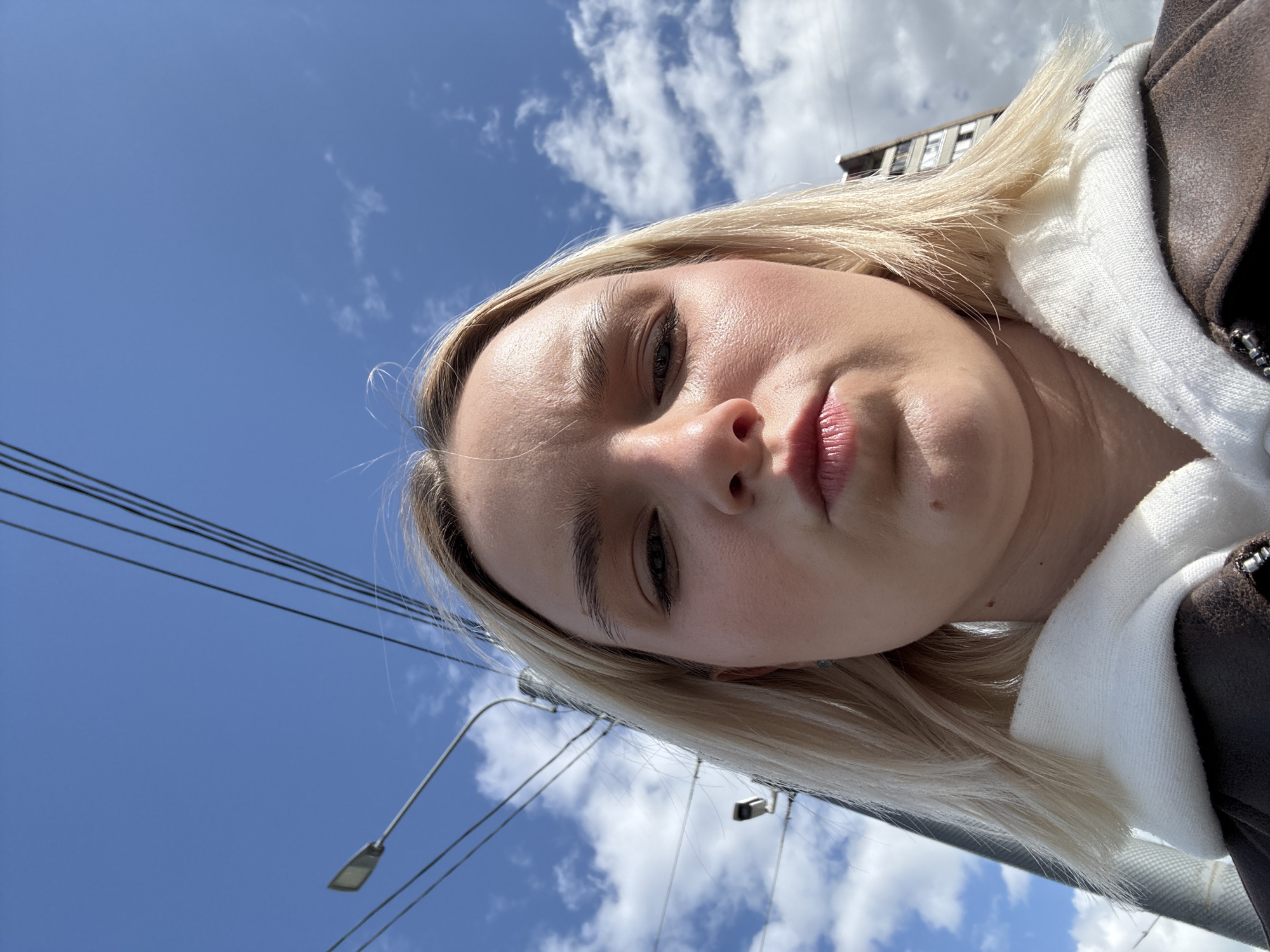 The image shows a selfie taken from a low angle, featuring a fair-skinned person with blonde hair. The individual has light eyebrows and appears to be wearing light makeup. They are wearing a white hooded sweatshirt under a dark brown jacket.
The background includes a bright blue sky with scattered white clouds. Electrical wires are visible crossing the upper left corner of the frame. There are street lamps in the background, along with a partially visible building.
Based on the architectural style and the presence of specific infrastructure, the location appears to be Saint Petersburg, Russia. The setting seems to be outdoors, likely a street or public area within the city.