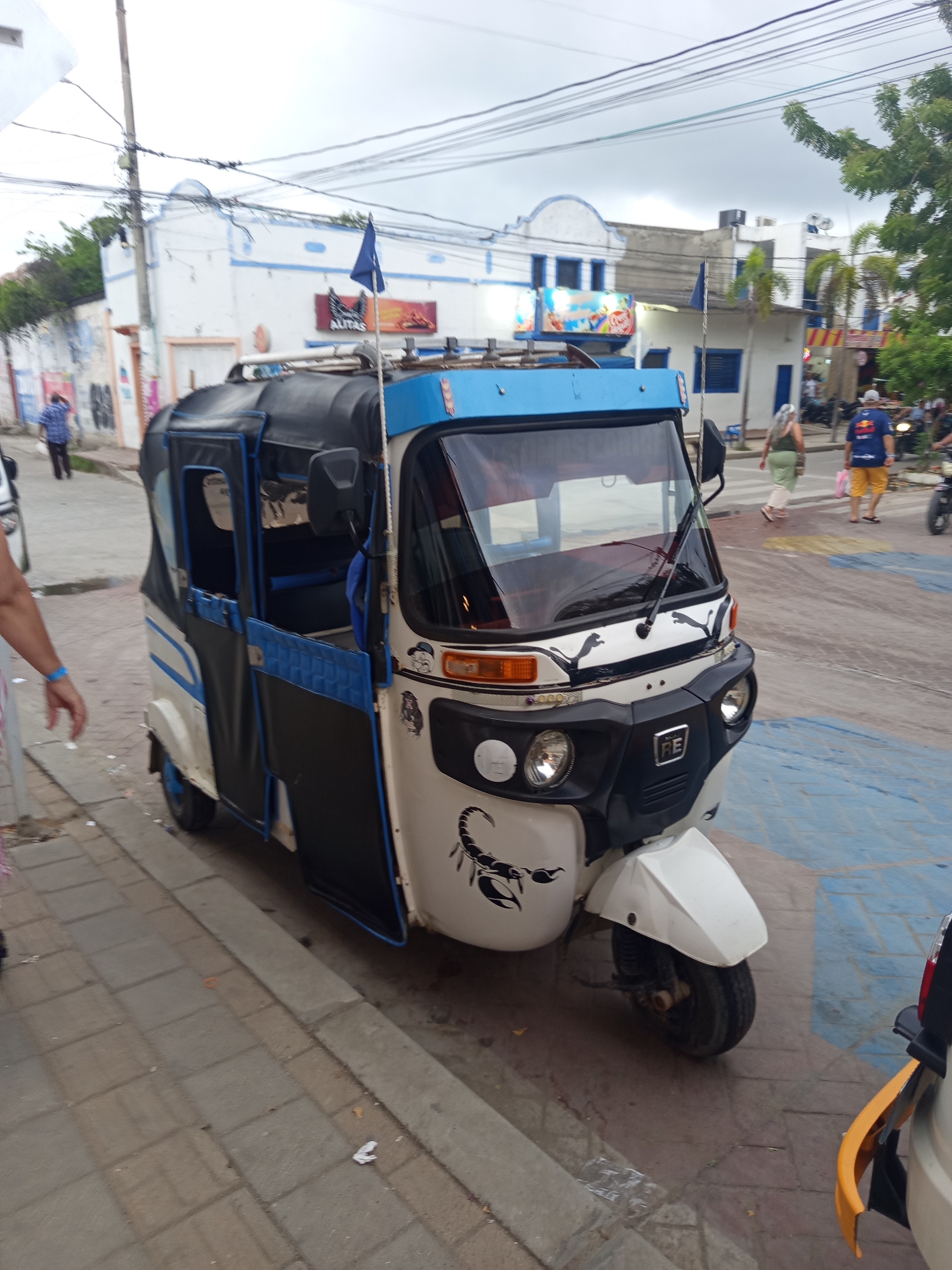 The image shows a white and blue three-wheeled auto rickshaw parked on a sidewalk in Tolú, Colombia. The auto rickshaw has black doors with blue trim and a blue canopy. A black scorpion decal is visible on the side of the vehicle. In the background, there are white buildings with blue trim, power lines, and some trees. Several people are walking in the background, and a motorcycle is parked on the right side of the image. The sky is overcast. The sidewalk is made of stone pavers.