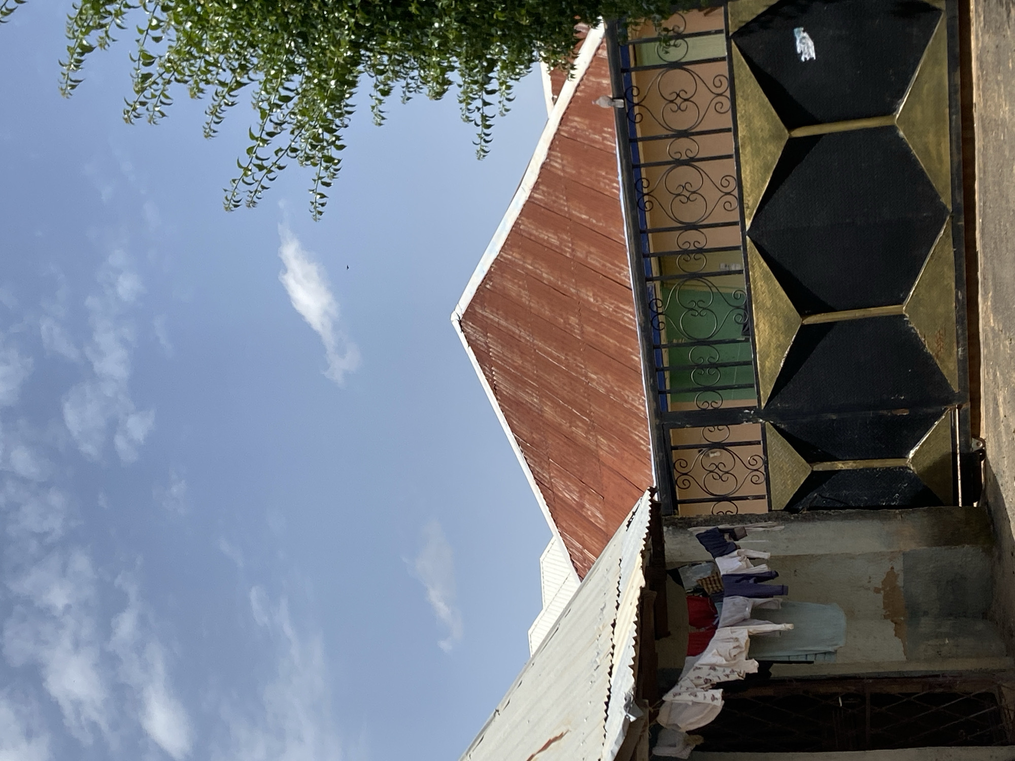 The image captures a scene in Bar Jirgi Summa, Nigeria, on a bright, sunny day with a blue sky dotted with scattered white clouds. In the foreground to the left, laundry hangs on a clothesline attached to a weathered, light-colored wall. The laundry includes several white and patterned garments. To the right of the laundry, a black metal gate with gold triangular accents and decorative scrollwork separates the viewer from a courtyard or street. Behind the gate, a building with a pale orange facade is visible, featuring green-colored window frames. Dominating the mid-ground are several pitched roofs. The most prominent is a large, rusty-red metal roof with a white trim. To its left, a smaller, corrugated metal roof of a lighter, weathered silver color partially obscures another white roof behind it. The scene is framed on the right by the lush green foliage of a tree. A small bird is visible in flight in the sky, and a pigeon is perched on the fence above the gate.