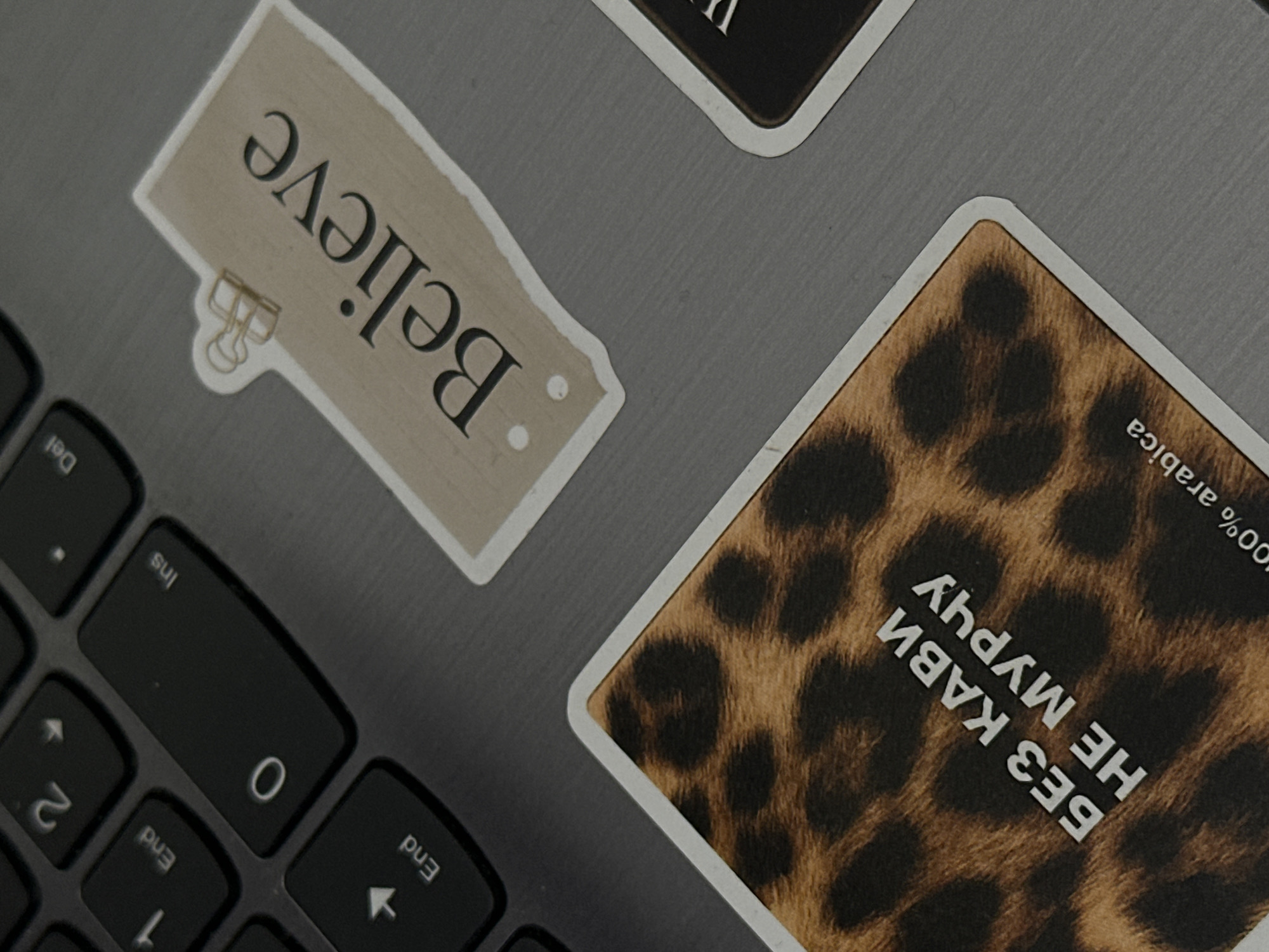 The image shows a close-up view of a laptop keyboard with several stickers. One sticker has the word "Believe" printed on it in a serif font, with a small golden paperclip holding it in place. Another sticker features a leopard print pattern with text in Cyrillic that translates to "Without coffee, no meow." Below that, the text "100% arabica" is visible. There is also a partial view of a third sticker with dark text. The keyboard itself is dark gray and shows keys like "End," "0," "Ins," and "Del." The lighting suggests an indoor environment, possibly a desk. The image does not provide any information about the city of Desna, Ukraine.