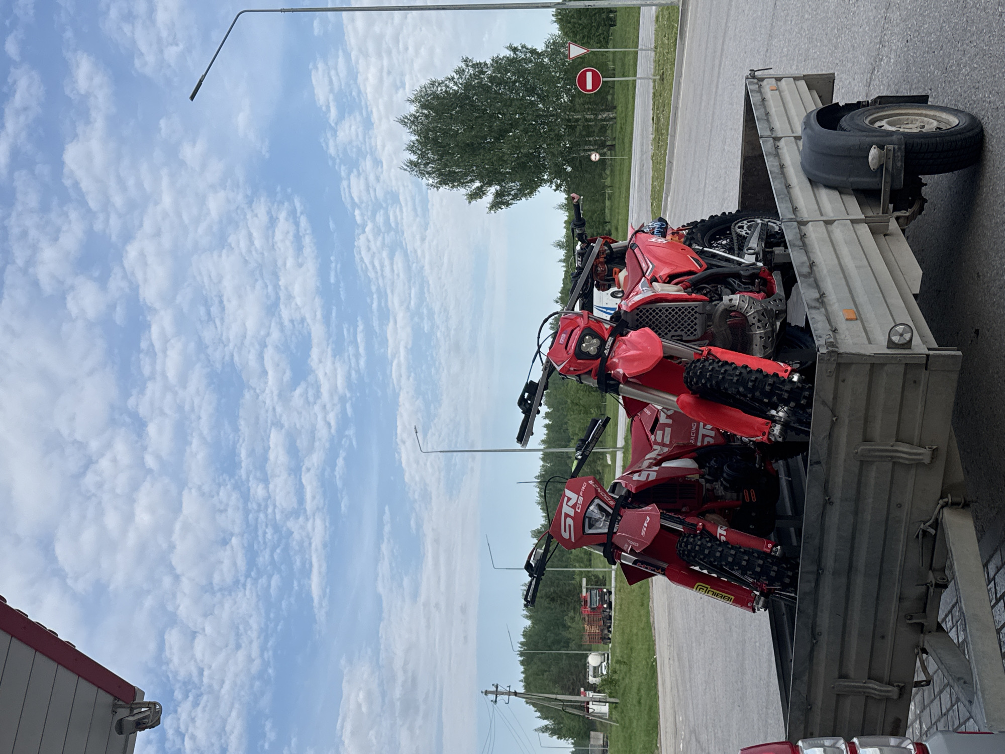 The image shows three red dirt bikes loaded onto a grey metal trailer. The trailer is parked on the side of a road. A spare tire is attached to the front of the trailer. There is a "no entry" road sign visible in the background. Trees and utility poles are visible in the background. The sky is blue with white clouds. Based on the metadata, the location is Marchugi, Russia.