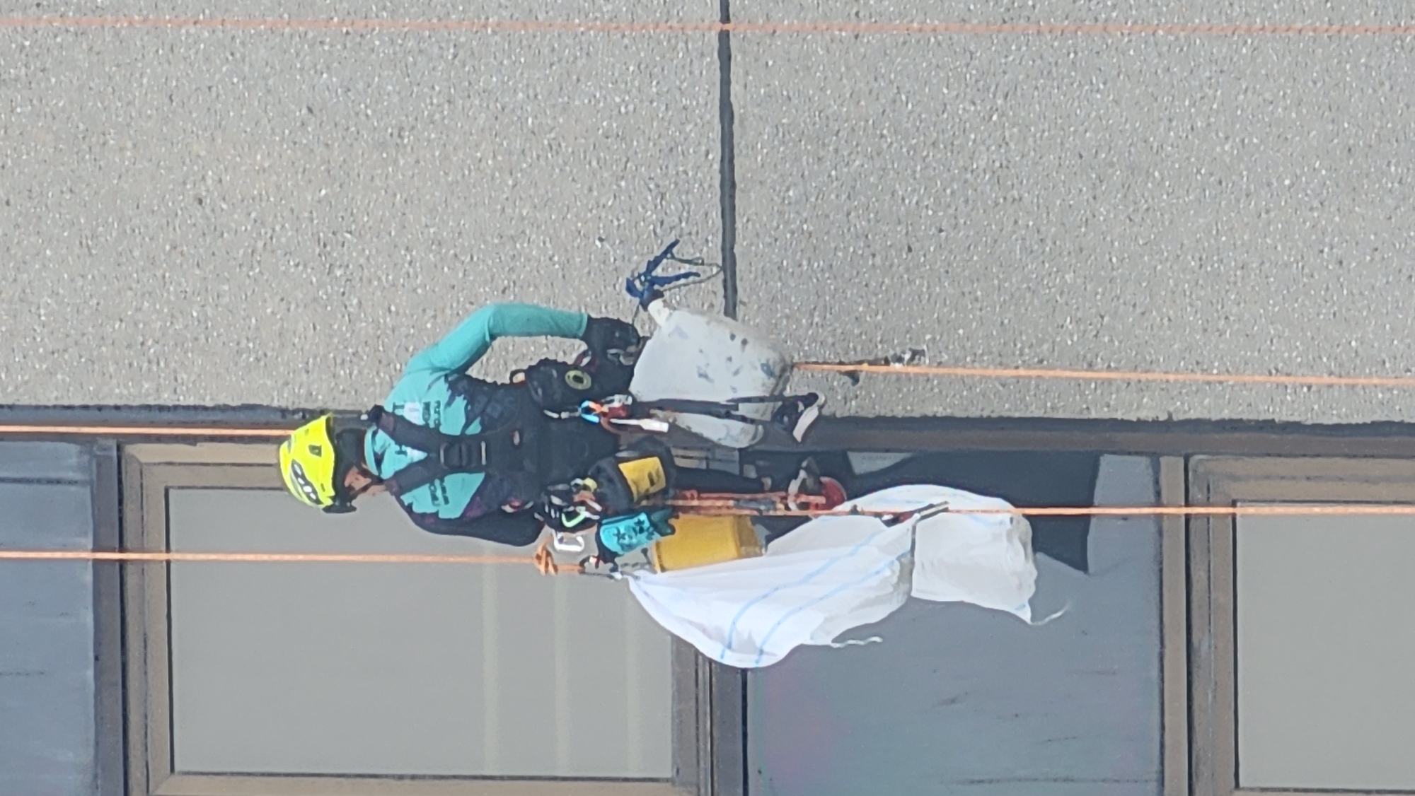 A person is rappelling down the side of a building, wearing a yellow helmet and a turquoise and black climbing harness. They are suspended by ropes and are carrying several bags and buckets, including a yellow bucket and a white bag. The building appears to be made of concrete, and there is a window on the left side of the image. The scene is outdoors, and the lighting suggests it is daytime. There is no information in the image to suggest the location is Moscow, Russia.