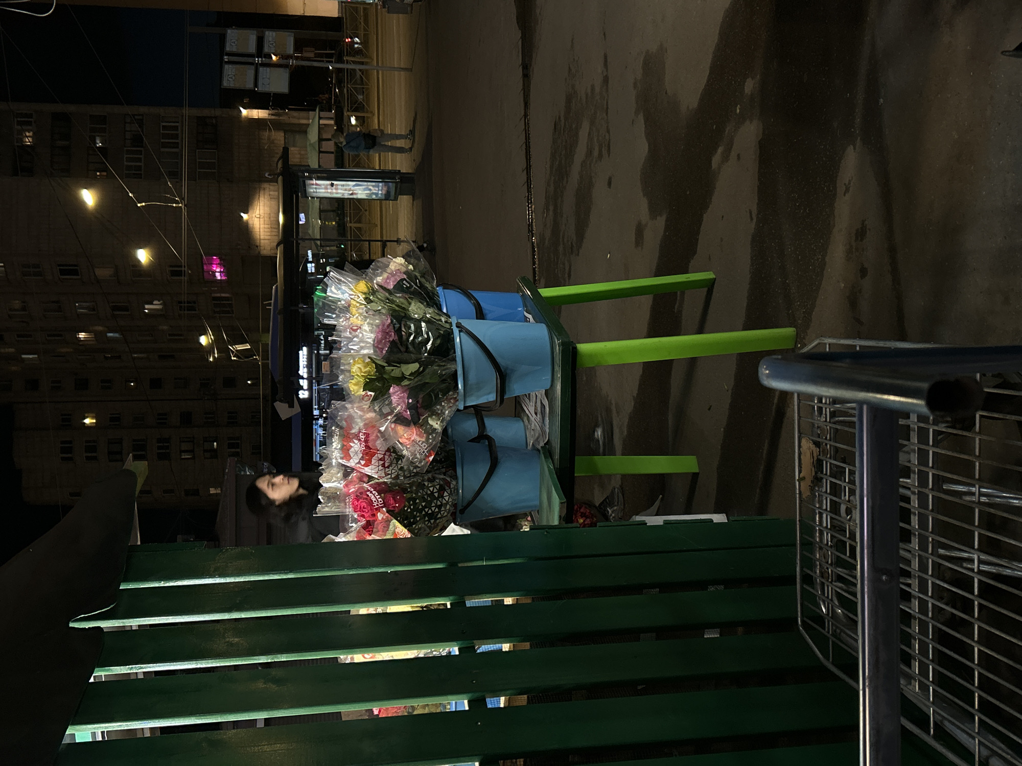 The image depicts an outdoor scene in Saint Petersburg, Russia, at night. A woman stands near a flower stand, which holds buckets of flowers wrapped in plastic. The flowers are a variety of colors including yellow, pink, and red. A green wooden bench sits in the foreground. A shopping cart is partially visible on the right side. In the background, a multi-story building is seen, lit by streetlights. A pedestrian can be seen in the distance. The ground appears wet.