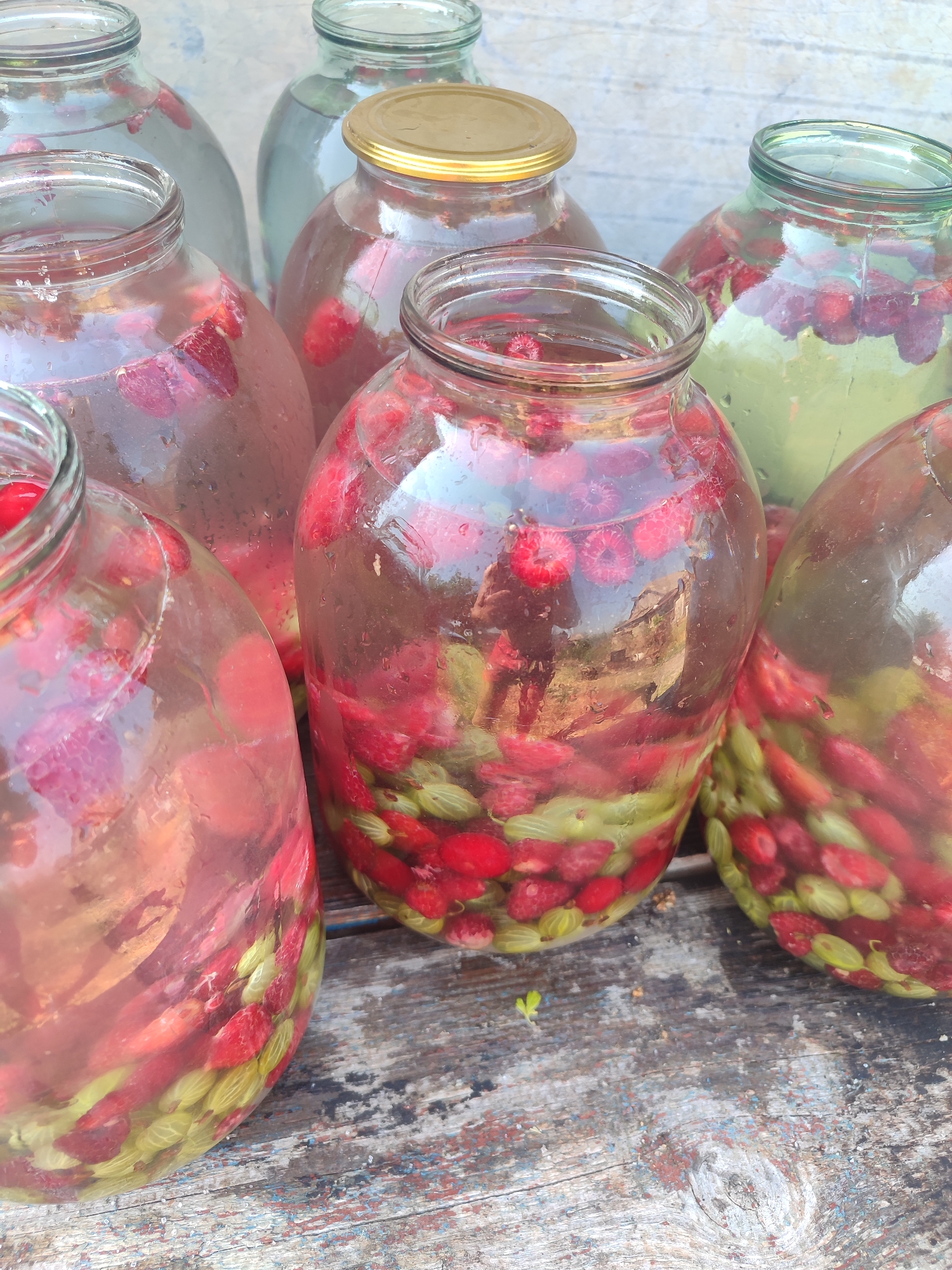 An outdoor, close-up, eye-level, vertical shot shows several large glass jars filled with raspberries and possibly gooseberries, submerged in a clear liquid. The jars are arranged on a weathered wooden surface. In the foreground, two jars are most prominent, their contents clearly visible. One jar in the center reflects a person taking the photo. In the background, more jars are partially visible. The overall impression is one of homemade preservation. The location is identified as the city of Novosamarka, Ukraine.
