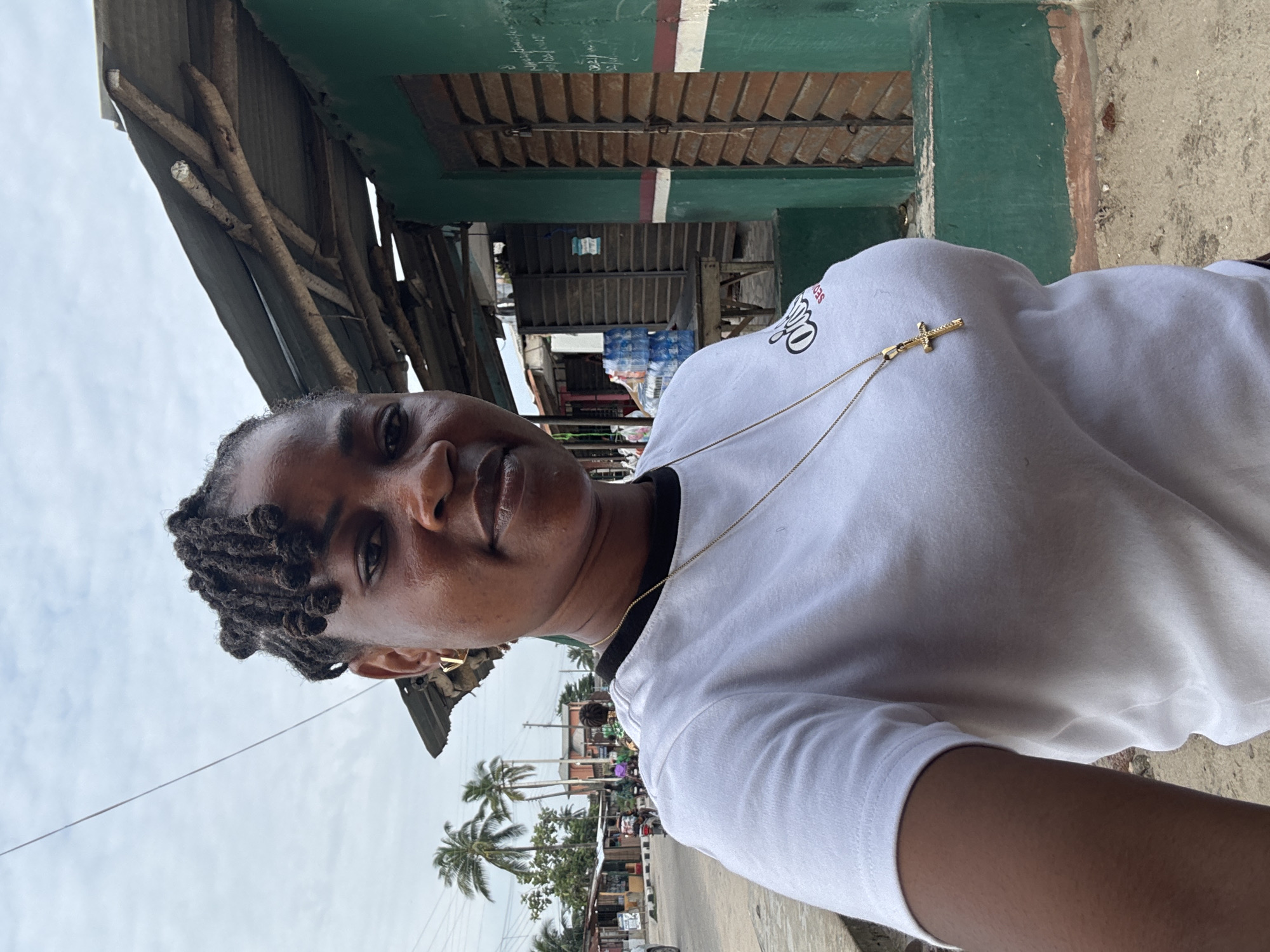 A woman, with dark skin and braided hair, stands in front of a green building with shuttered windows. She is wearing a white t-shirt with a black collar and a gold necklace with a cross pendant. In the background, there are palm trees, a street with traffic, and other buildings. The scene appears to be in a market area. The sky is overcast.