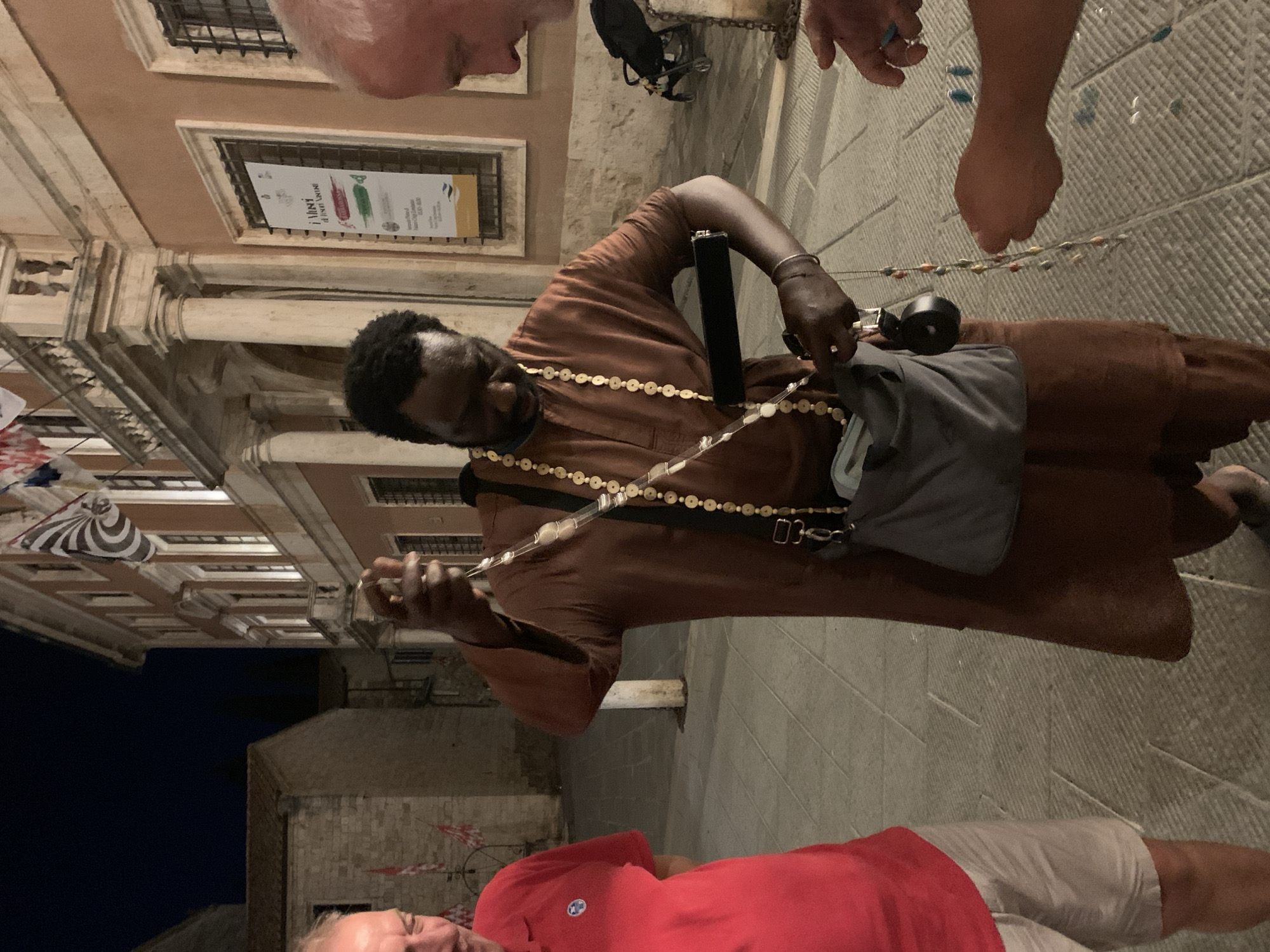 A man in a brown robe is selling beaded necklaces on a cobblestone street in San Quirico d'Orcia, Italy, at night. He is holding a necklace up for a potential customer to see, while a third man in a red shirt and shorts watches. In the background, a building with a balcony and flags is illuminated by lights.