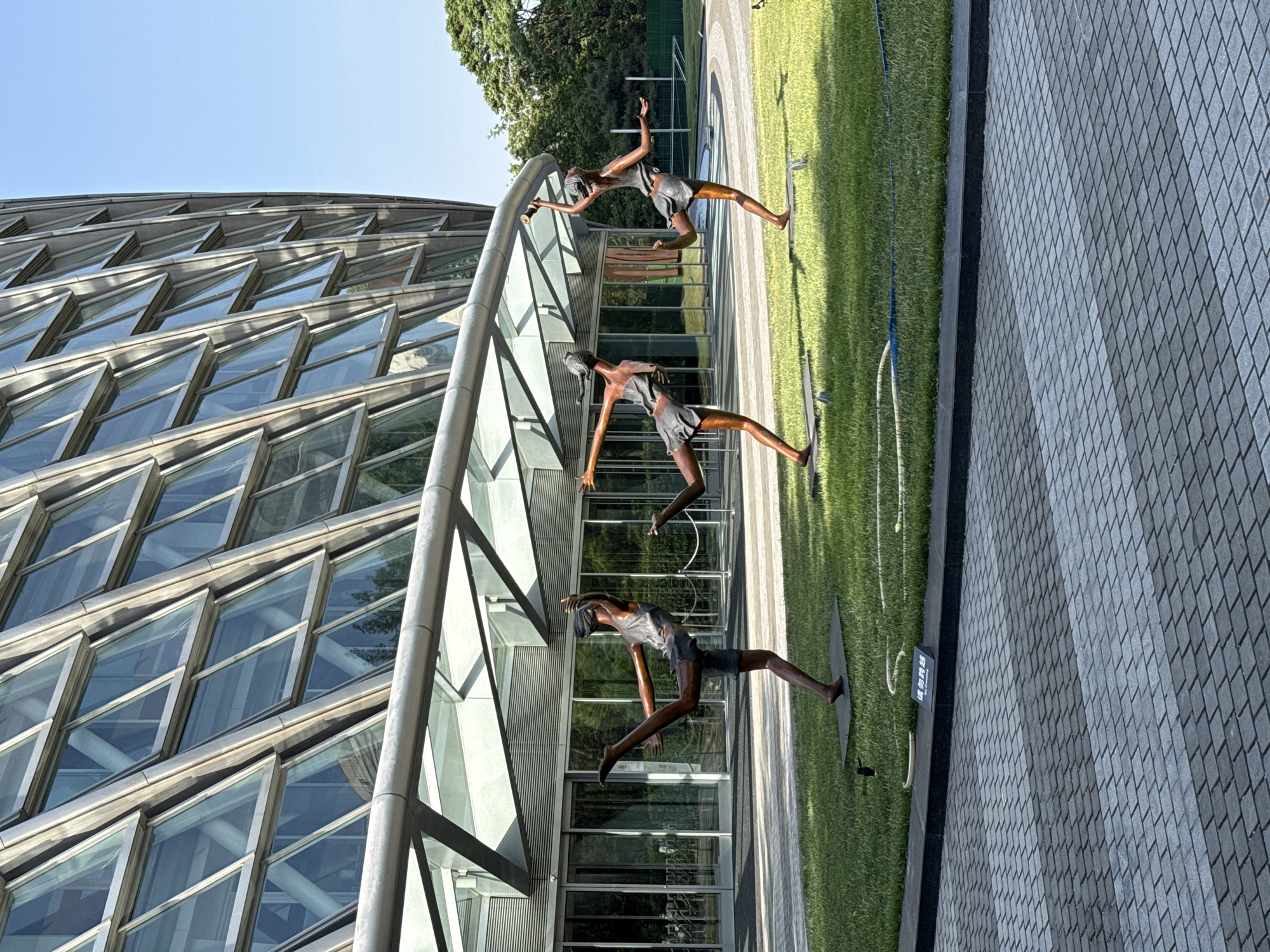The image shows an outdoor sculpture in Chaoyang District, China. The sculpture depicts three figures in motion, possibly dancers or athletes, made of a dark metal, possibly bronze, arranged along a curved, modern architectural structure with a glass facade. The background includes a building with a distinctive glass and steel exterior, resembling a series of triangular windows, and a patch of green grass. A gray paved area is in the foreground.