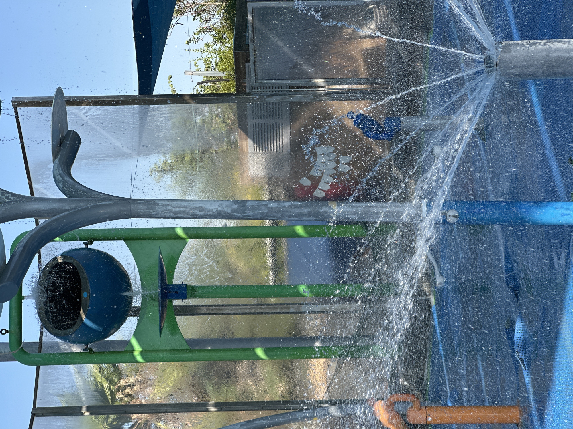 An outdoor water park feature in Hod HaSharon, Israel, shows a blue sphere-shaped bucket filling with water and beginning to spill. The bucket is suspended within a green frame structure. In the foreground, water sprays from a nozzle on the ground, creating a fan of water droplets. Other water features, including a blue pipe and an orange structure, are visible. A transparent panel is behind the water feature, showing a hint of greenery and buildings in the background. The ground is a blue surface.