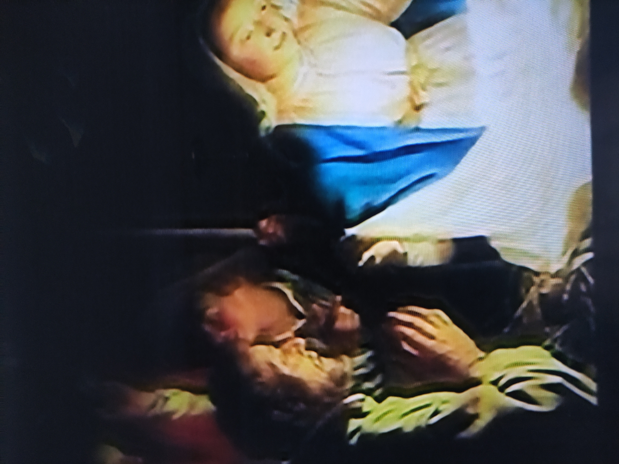 The image shows two individuals. One appears to be a blond woman lying down, dressed in a white top and blue pants or skirt. The second individual is a man with dark hair lying below her, seemingly resting on a green or yellowish surface. His face is visible, and his hands are near him. The environment is poorly lit, making it difficult to discern specific details. No information connects this image to Pavlodar, Kazakhstan.