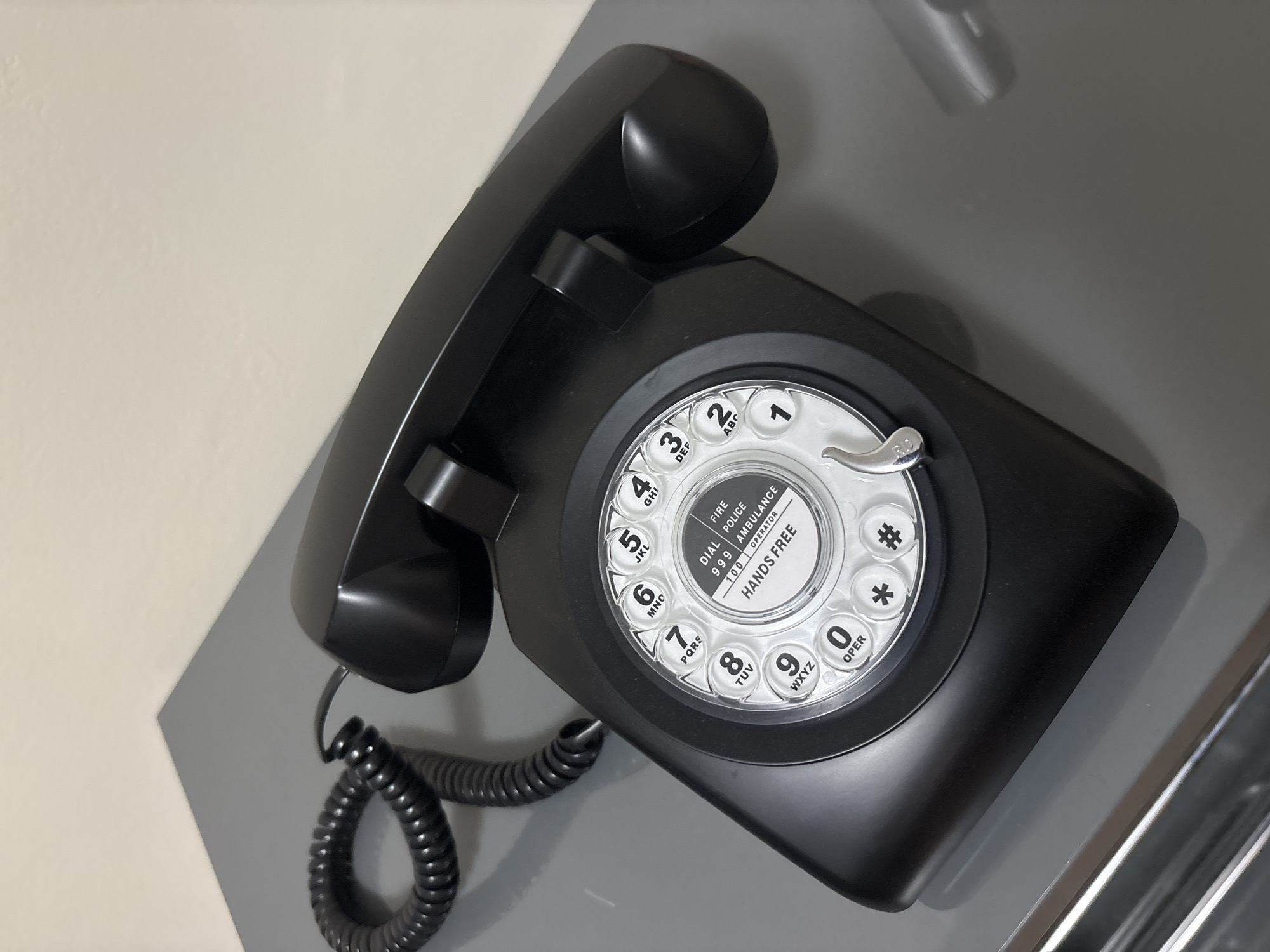 A close-up studio shot reveals a glossy black rotary telephone resting on a two-toned gray surface, with a pale gray wall in the upper left corner. The telephone's receiver is placed on the body of the phone, and the curled cord is visible on the left side. The rotary dial is white with black lettering and numbers, displaying emergency contact numbers. The photograph is taken from a slightly elevated angle.