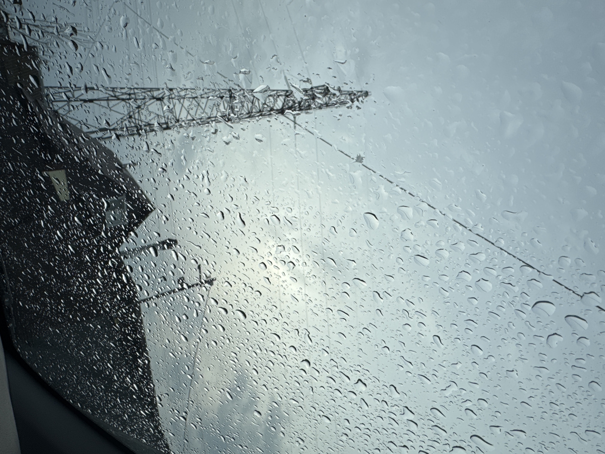 The image shows a rainy view through a window, with numerous water droplets covering the glass surface. In the background, there is a blurred view of a crane, likely used for construction, against a cloudy sky. A dark building is visible on the left side of the frame. The location is potentially in or near Chaikovsky, Russia, based on the provided information. There are no people visible in the image.