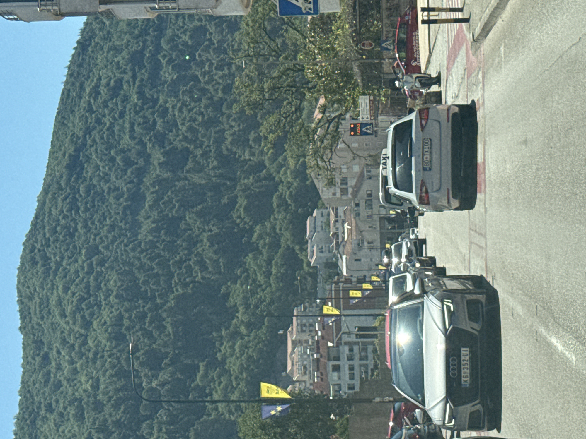 A grey Audi A6 sedan with license plate KG 6352-UI is in the foreground of the image, driving on a street in Budva, Montenegro. The car is facing forward, and its headlights are on. Behind the Audi, there is a white taxi van and other vehicles. In the background, there are buildings and a lush green mountain. The sky is blue and clear. There are several yellow flags with blue stars hanging from poles on the side of the road. A traffic light is visible in the distance, showing red.