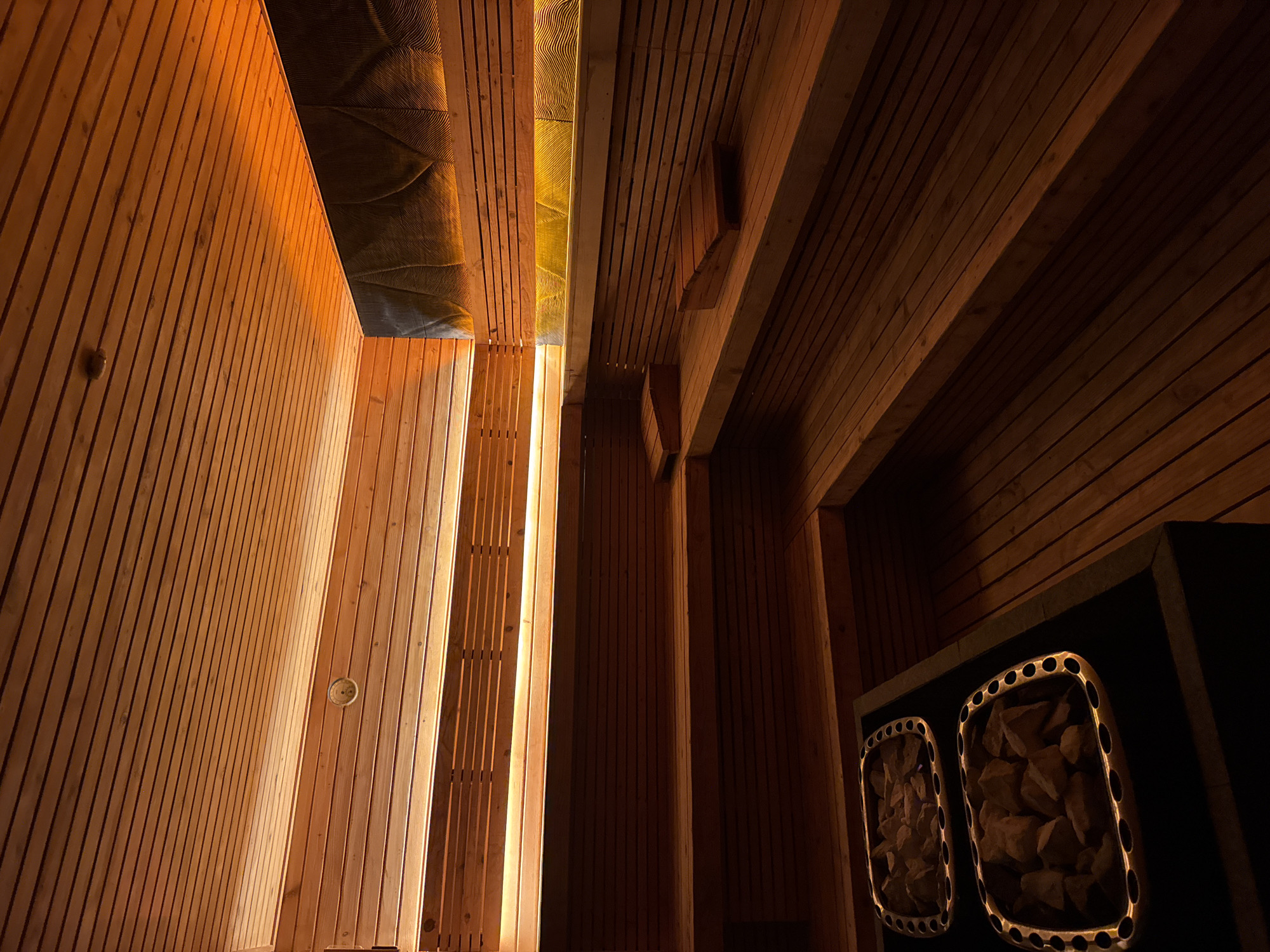 The image depicts the interior of a sauna. The walls and ceiling are constructed from light-colored wood planks. The sauna contains a heater with two compartments filled with rocks and a textured ceiling. There is an electrical outlet on the wall and the lighting creates a warm ambiance. The location of this sauna is unknown, as there are no identifying marks or features.