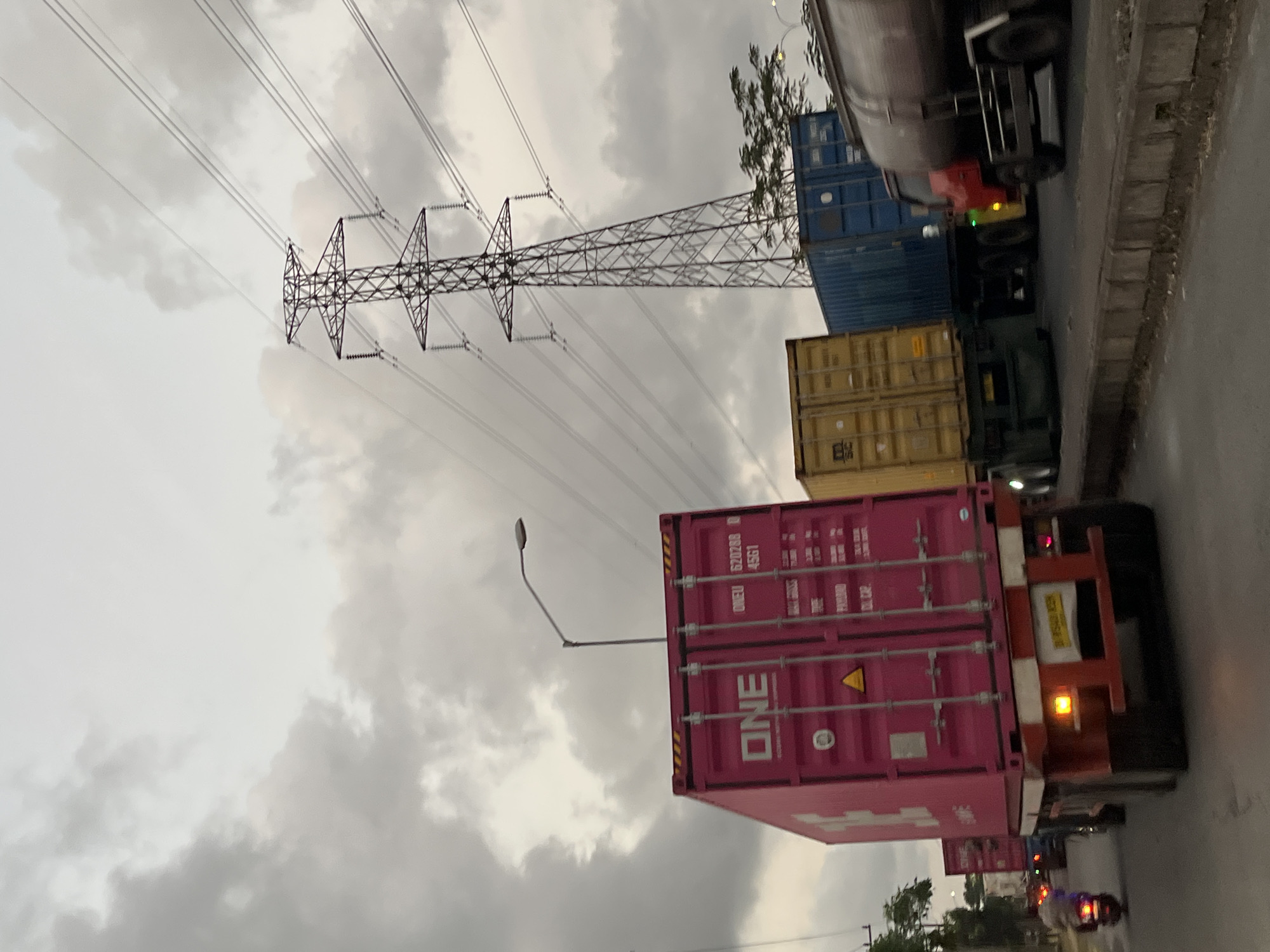 This image depicts a congested traffic scene on a road in Pamotan, Indonesia, under an overcast sky. In the foreground, a pink shipping container with the "ONE" logo is prominently displayed on the back of a truck. The container has markings indicating its serial number "620288 45G1". To the right of the pink container, a yellow shipping container is visible on another truck, followed by a blue shipping container on a third truck. Further to the right, a large tanker truck is present. In the background, a tall electricity pylon with multiple power lines stretches across the sky. A street lamp is also visible to the left of the pink container. Traffic appears to be moving slowly or stopped. A motorcyclist is seen in the distance, to the left of the pink container, heading in the same direction as the trucks. The road is lined with a low concrete barrier. The overall environment suggests an industrial or port area, given the presence of shipping containers.