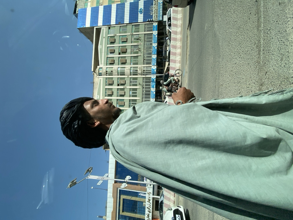 The image depicts a street scene in Kandahar, Afghanistan, on a clear, sunny day. In the foreground, viewed from the right side, is a man wearing a dark, coiled turban and a light teal-green shawl draped over his left shoulder and body. He has a beard and mustache, and his gaze is directed slightly downwards and to the right. His right arm is visible, featuring a silver-colored wrist bracelet and a small, light-colored object held in his hand.
Behind the man, a multi-lane asphalt road stretches horizontally across the midground. A red and white striped concrete barrier separates lanes. Vehicle traffic is present; a white sedan is partially visible on the far left, and a motorcycle carrying two individuals in light-colored clothing is moving from right to left beyond the barrier. Other vehicles are faintly visible further down the street.
In the background, several multi-story buildings line the street under a bright blue sky. On the left, a light-colored building with blue accents displays signage with what appears to be Dari or Pashto script. To its right, a prominent light green building features multiple windows, many equipped with small, light-colored louvered shutters. Adjacent to the green building on the far right, another building is partially visible with a distinctive blue and white striped facade. Streetlights with multiple fixtures extend upwards on the left side of the frame. Reflections are present on the upper portion of the image, suggesting the photograph was taken through glass.
