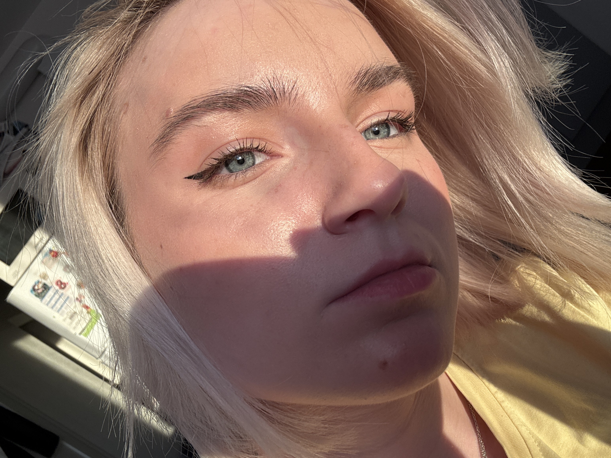 A close-up, angled portrait of a fair-skinned person with blonde hair and blue eyes. The subject is wearing winged eyeliner. The lighting is bright, casting shadows across the face. The person is wearing a yellow top and a silver necklace is visible. The location is Unknown, Unknown.