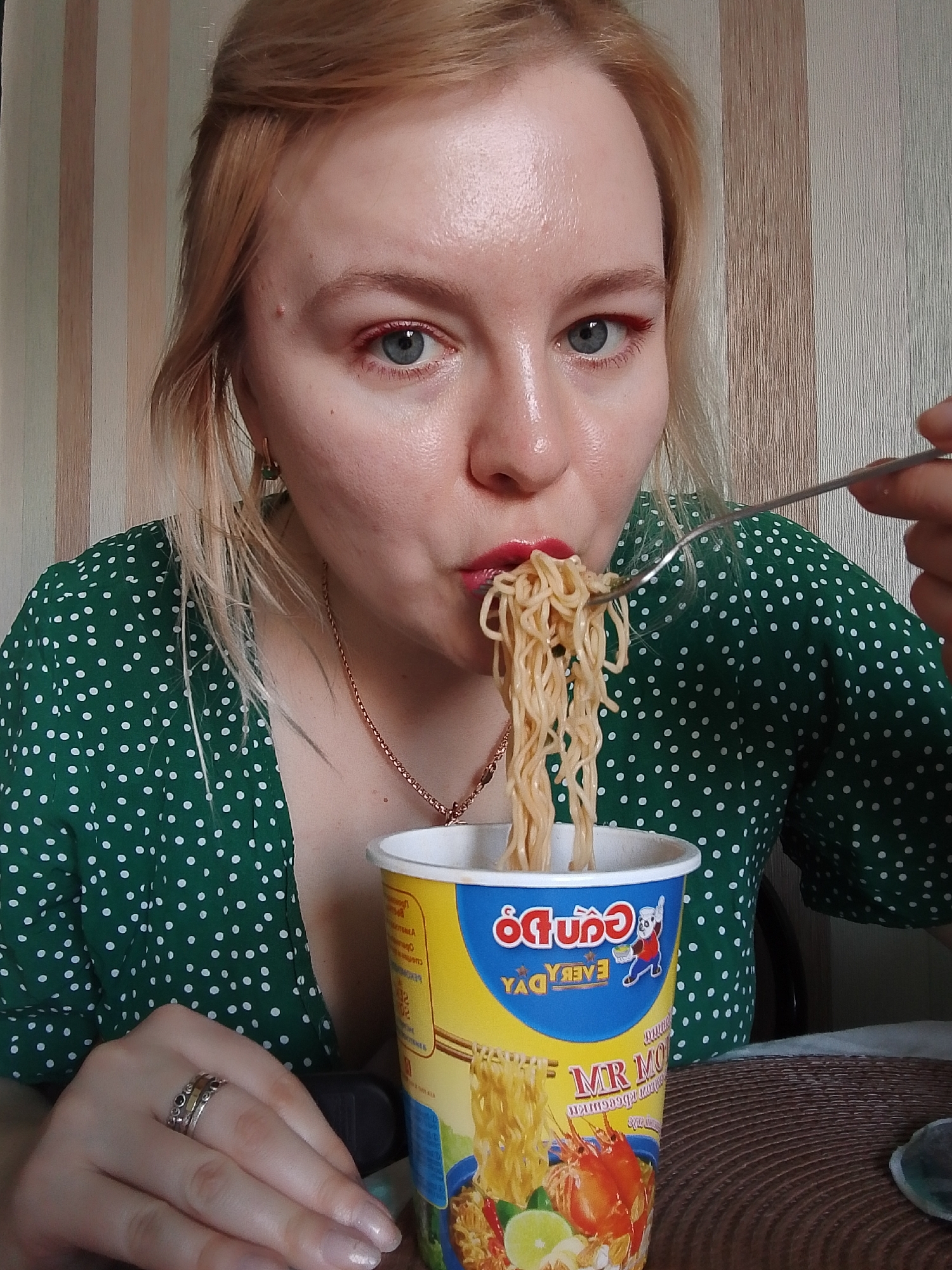 A young woman with fair skin and blonde hair is eating noodles from a yellow and blue cup labeled "Cuti Do EVERY DAY MR MO", possibly instant noodles, with a fork. She is wearing a green, white-dotted dress and a gold necklace and has a ring on her finger. She has red lipstick and possibly red eyeliner. The background appears to be an interior setting, potentially a home. The location in Izhevsk, Russia is not discernible from the image.