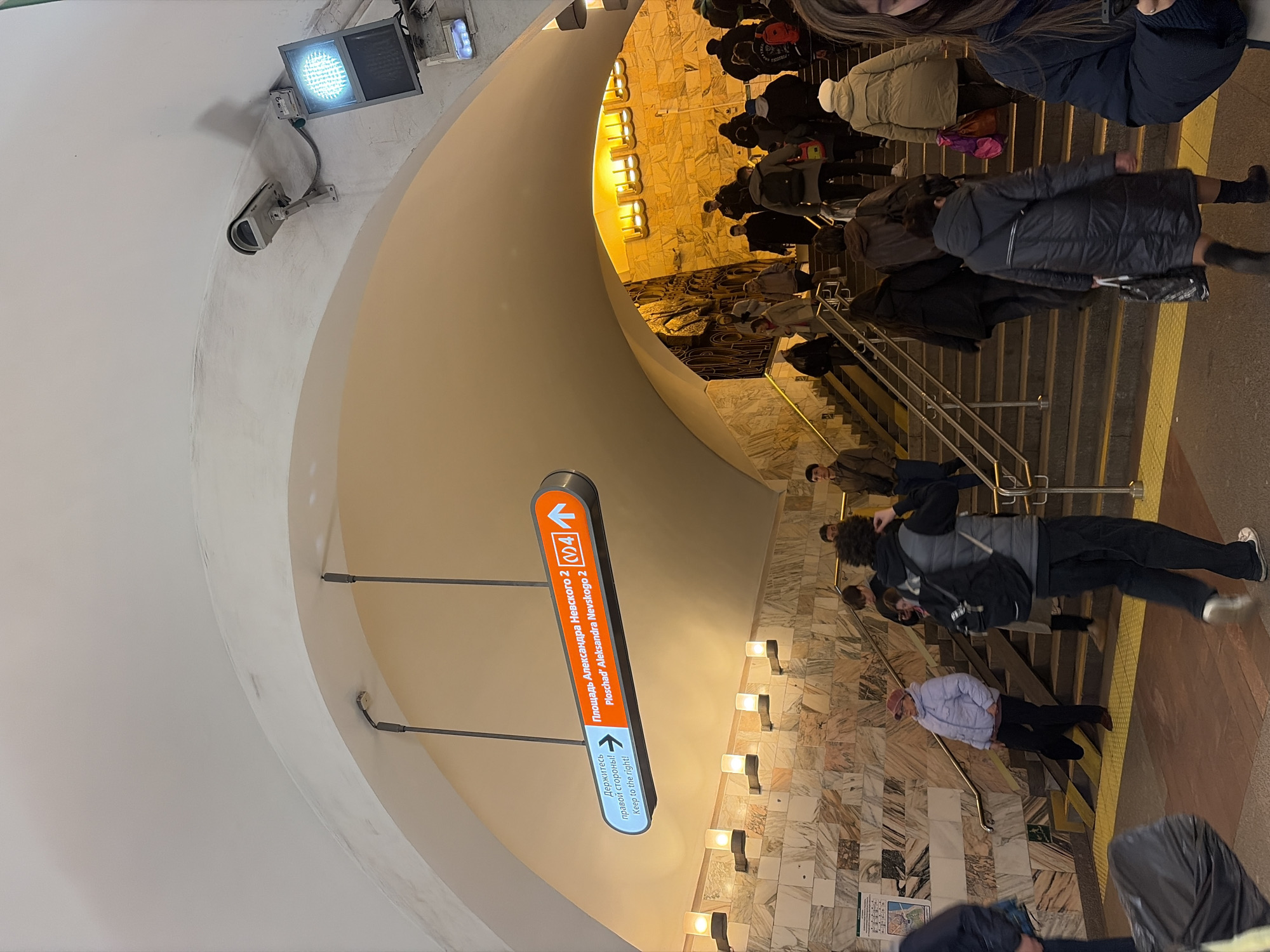 The image depicts a scene inside a metro station in Saint Petersburg, Russia. Many people are walking down a set of stairs. A direction sign, partially rotated, indicates the way to "Ploschad' Alexandra Nevskogo 2" (Alexander Nevsky Square 2), with an instruction to keep to the right. The walls are marble. A blue light fixture and a camera are mounted to the ceiling. The interior design of the metro station features a curved architecture with lighting and decorative elements.