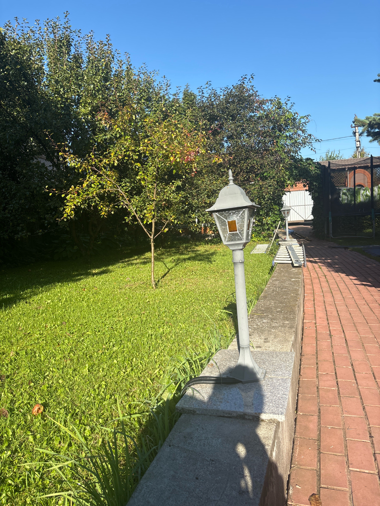 In Zhavoronki, Russia, a bright, sunny day illuminates a residential yard. The foreground features a gray, ornate lamppost casting a distinct shadow on a granite and concrete border. Adjacent to this border, a vibrantly green lawn stretches towards the middle ground, where a young tree with sparse foliage stands. Beyond the lawn, dense trees with dark green and reddish-brown leaves form a lush backdrop, suggesting the transition from summer to autumn. To the right of the border, a red brick pathway guides the eye into the distance, where a white double gate and a dark metal fence are visible. The sky above is a clear, deep blue.