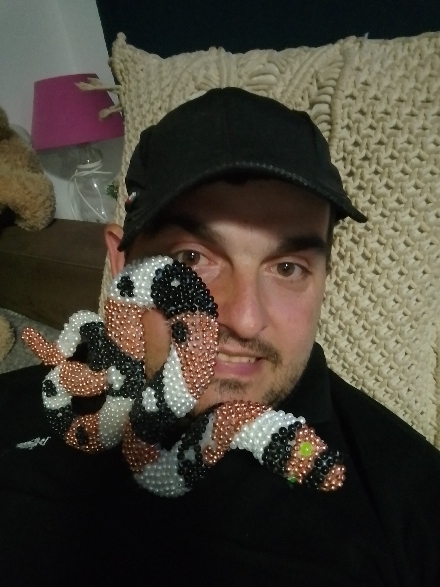 A close-up, medium shot captures a man wearing a black baseball cap and a black shirt. He is holding a beaded snake-like object, predominantly in white, black, and copper tones, near his face, partially obscuring his right eye and cheek. The snake has visible green bead eyes. The man has brown eyes, a light complexion, and stubble. Behind him, a textured, cream-colored blanket or throw is visible. To the left of the frame, a portion of a brown stuffed animal and a pink lampshade can be seen. The lighting suggests an indoor setting, likely a home environment.