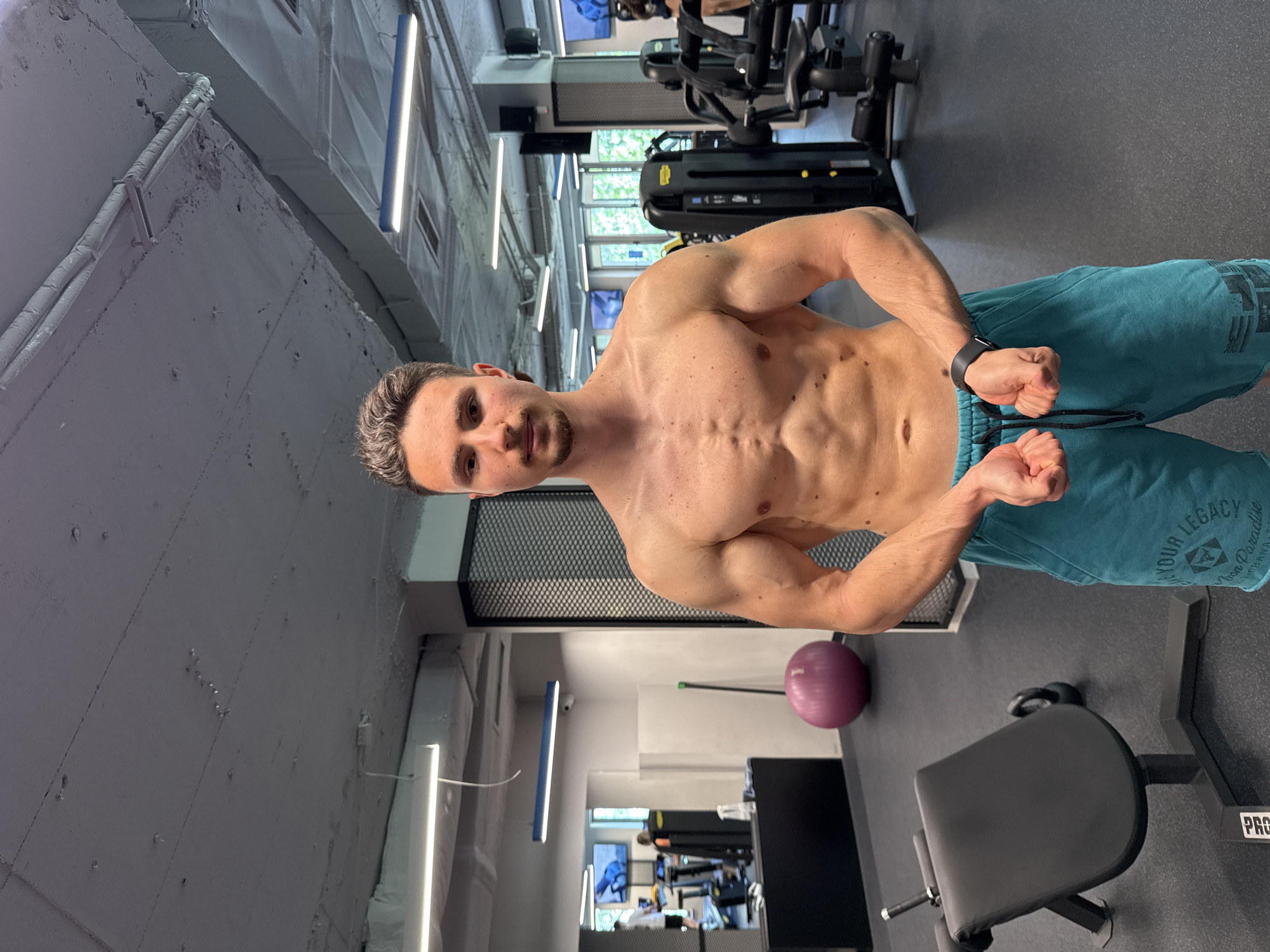 A shirtless man is striking a pose in a gym. He is muscular and wearing teal shorts with the words "YOUR LEGACY" printed on them. He has short brown hair and a light beard. The gym has workout equipment in the background and is indoors. The location appears to be in the city of Sofia, Bulgaria.