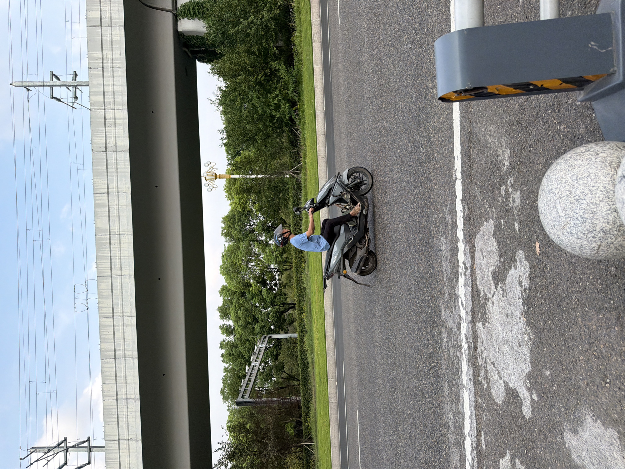 A person wearing a blue shirt and a helmet rides a gray scooter on a paved road. The scooter is moving from left to right in the frame. Behind the scooter, a green grassy area with trees is visible. Above the trees, a large, gray concrete overpass stretches horizontally across the image. Overhead, power lines and utility poles are suspended. The sky above is blue with some clouds. In the foreground, the road surface is visible with white lane markings. To the right of the frame, a gray barrier with yellow and black markings is present. The scene appears to be in an urban or suburban area of Jiangjin, China, given the infrastructure and vegetation.